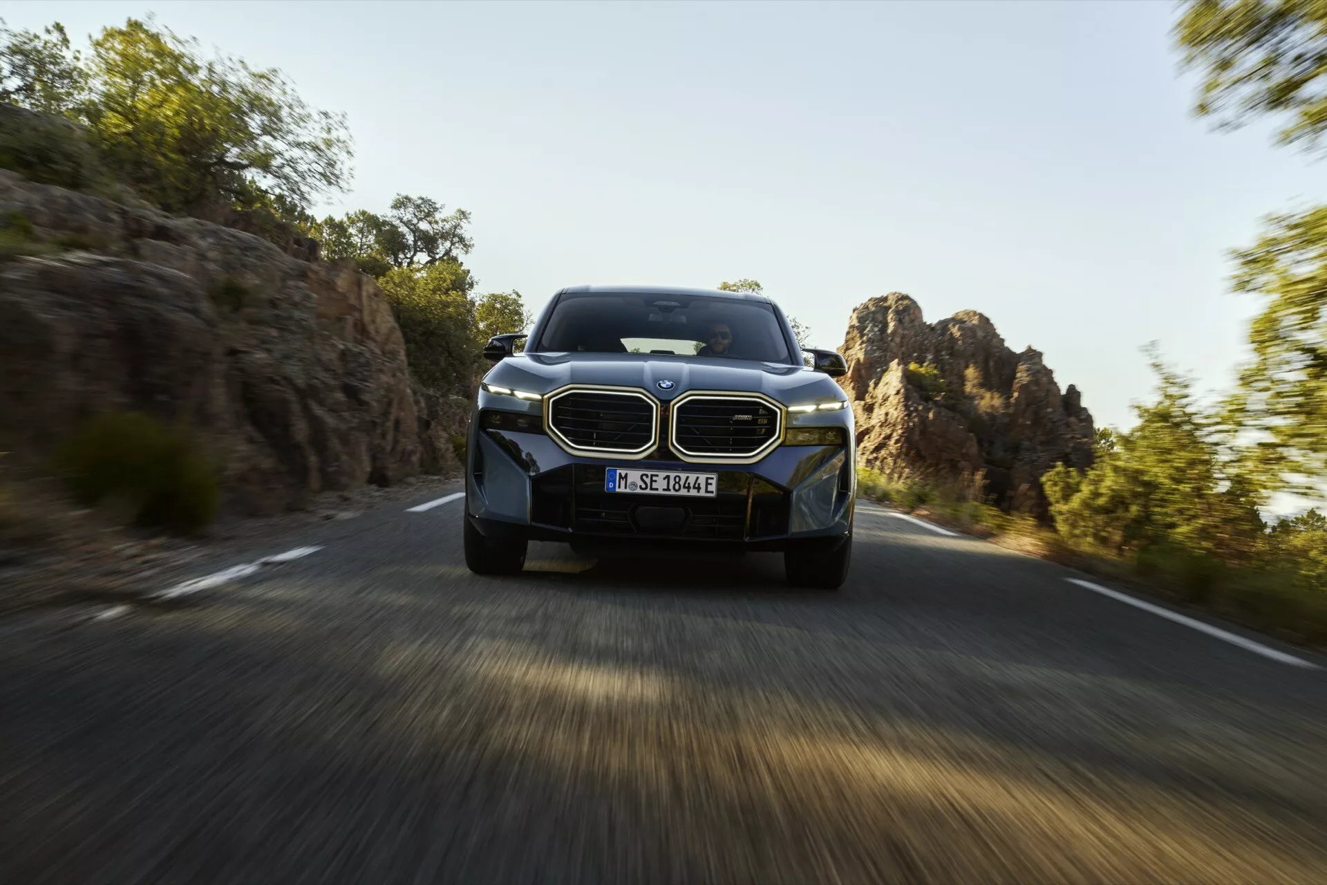 BMW XM 2023 (G09) sa pohybuje po kľukatej ceste s efektom rozmazania pohybu, ktorý ukazuje jeho veľkú obličkovitú mriežku, štíhle LED svetlomety a moderné zliatinové kolesá v prírodnom, skalnatom teréne.