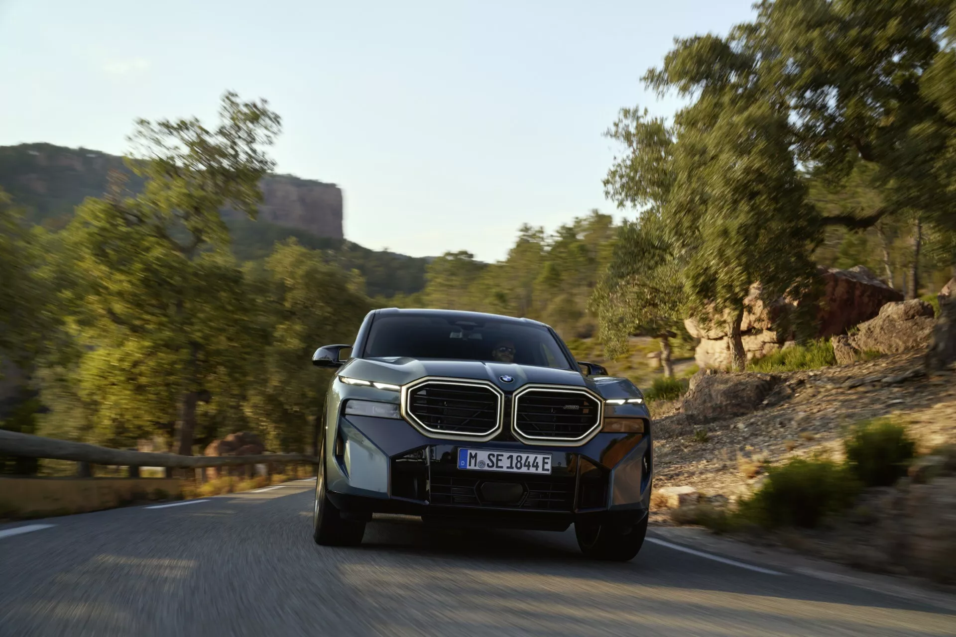 Predný pohľad na BMW XM 2023 (G09) v pohybe s dynamickým uhlom, ktorý zdôrazňuje jeho veľkú mriežku, LED svetlomety a elegantný dizajn, naznačujúci hybridnú technológiu, na vidieckej ceste