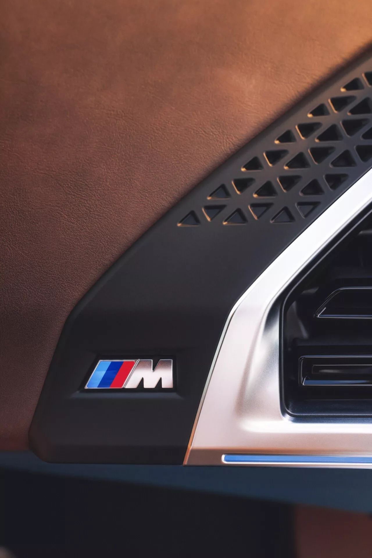 Detailný pohľad na interiér BMW XM 2023 (G09), zobrazujúci kožené čalúnenie, prvok so geometrickým vzorom, emblém M so symbolickými pruhmi a detail kovovej ventilácie