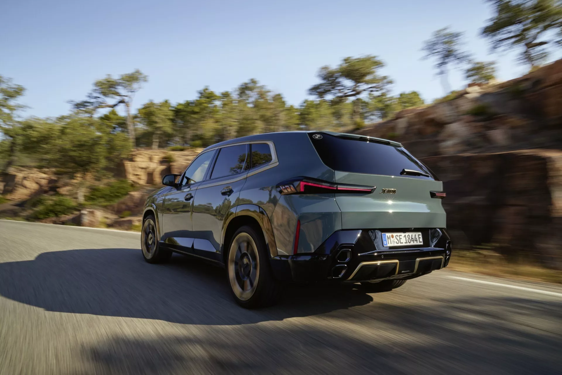 BMW XM (G09) z roku 2023 zachytené v dynamickom pohybe z pohľadu zozadu a z boku, čo zdôrazňuje jeho elegantný dizajn s LED zadnými svetlami, metalickým povrchom a kolieskami pre vysoký výkon na slnečnej ceste.