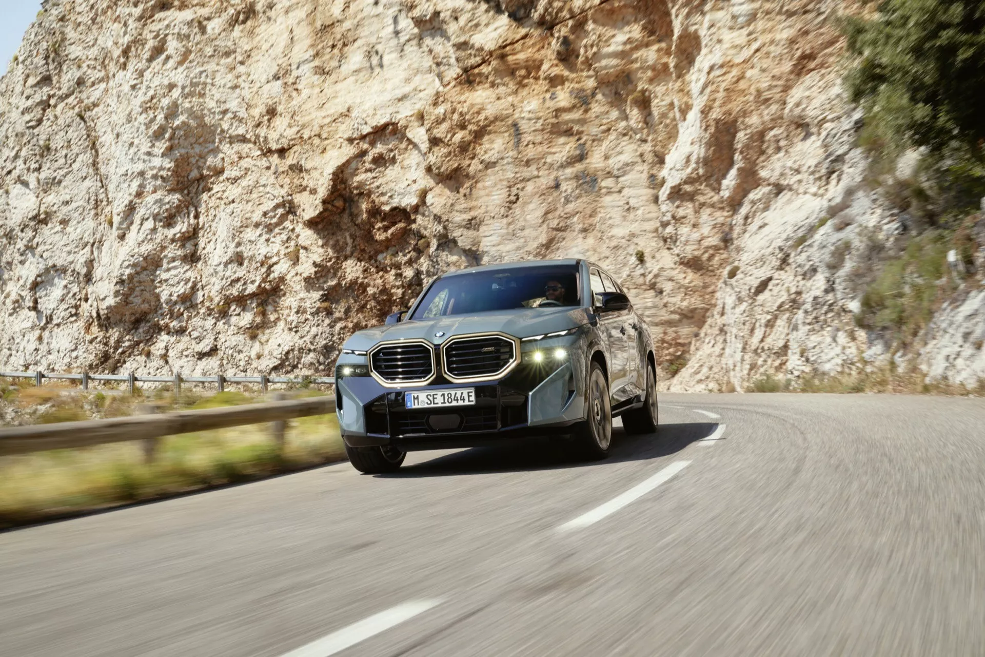 BMW XM (G09) z roku 2023 v dynamickom pohybe na zákrutej ceste, ukazuje jeho luxusný profil z predného trojštvrťového uhla, charakteristickú dvojitú obličkovitú mriežku, štíhle LED svetlomety, viacramenné zliatinové kolesá a aerodynamickú karosériu s metalickým lakom a chrómovými prvkami.