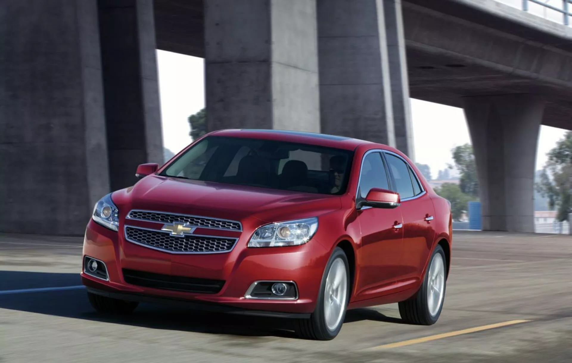 Dynamický pohľad na červený sedan Chevrolet Malibu 2012 v pohybe na moste, zobrazujúci predný dizajn s chrómovou mriežkou, štíhlymi svetlometmi, športovým siluetom a zliatinovými kolesami.