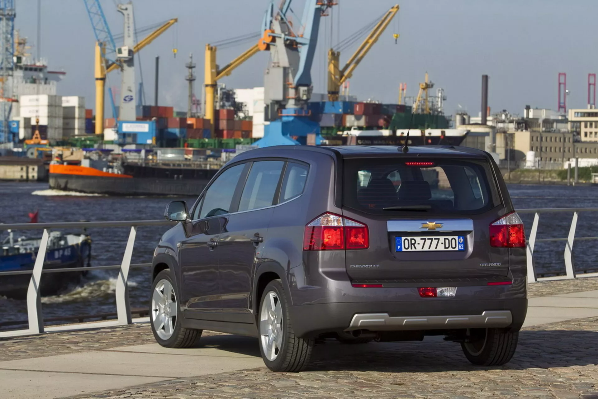 Zadný trojštvrťový pohľad na Chevrolet Orlando 2011 na nábreží prístavu, zdôrazňujúci jeho karosériu kompaktného SUV, päťdverovú konfiguráciu, dvojitý výfukový systém, senzory asistencie parkovania na nárazníku a zliatinové kolesá s viacerými perlami