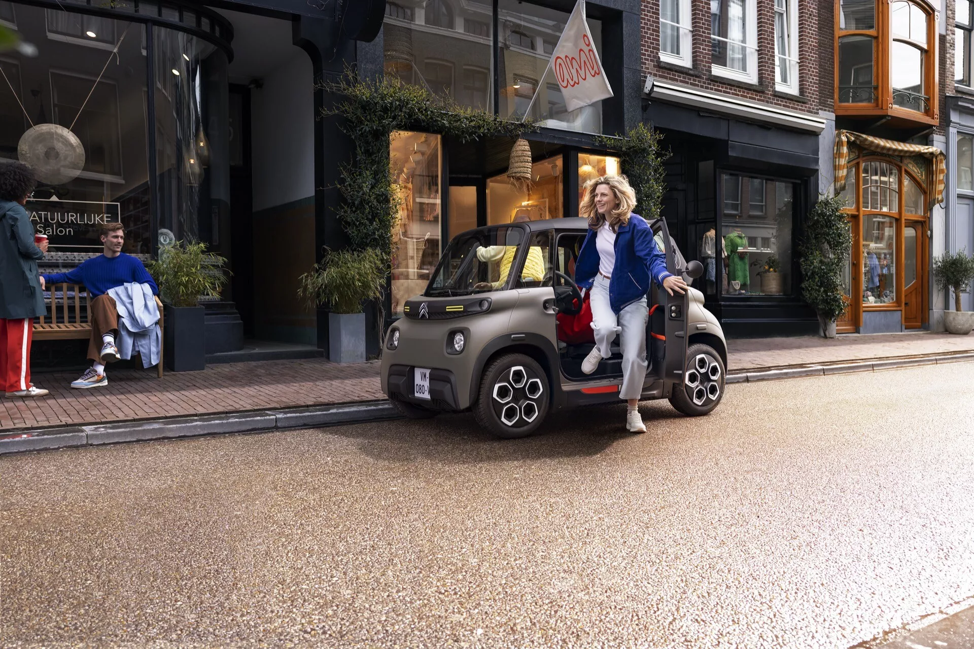 Na obrázku je Citroen Ami 2024 v približne trojštvrťovom uhle zpredu, ukazujúci jeho moderný a neobyčajný dizajn s okrúhlymi svetlometmi a vlnitým grilom, v mestskom prostredí s obchodmi v pozadí. Auto je kabriolet so stiahnutou strechou a bez dverí, čo umožňuje jasný pohľad na malé, viaclúčové kolesá a dvojfarebné ráfiky. Vedľa vozidla stojí neformálne oblečená osoba, ktorá dodáva prvok životného štýlu, zvýrazňujúc jeho mestský, ekologický obraz.
