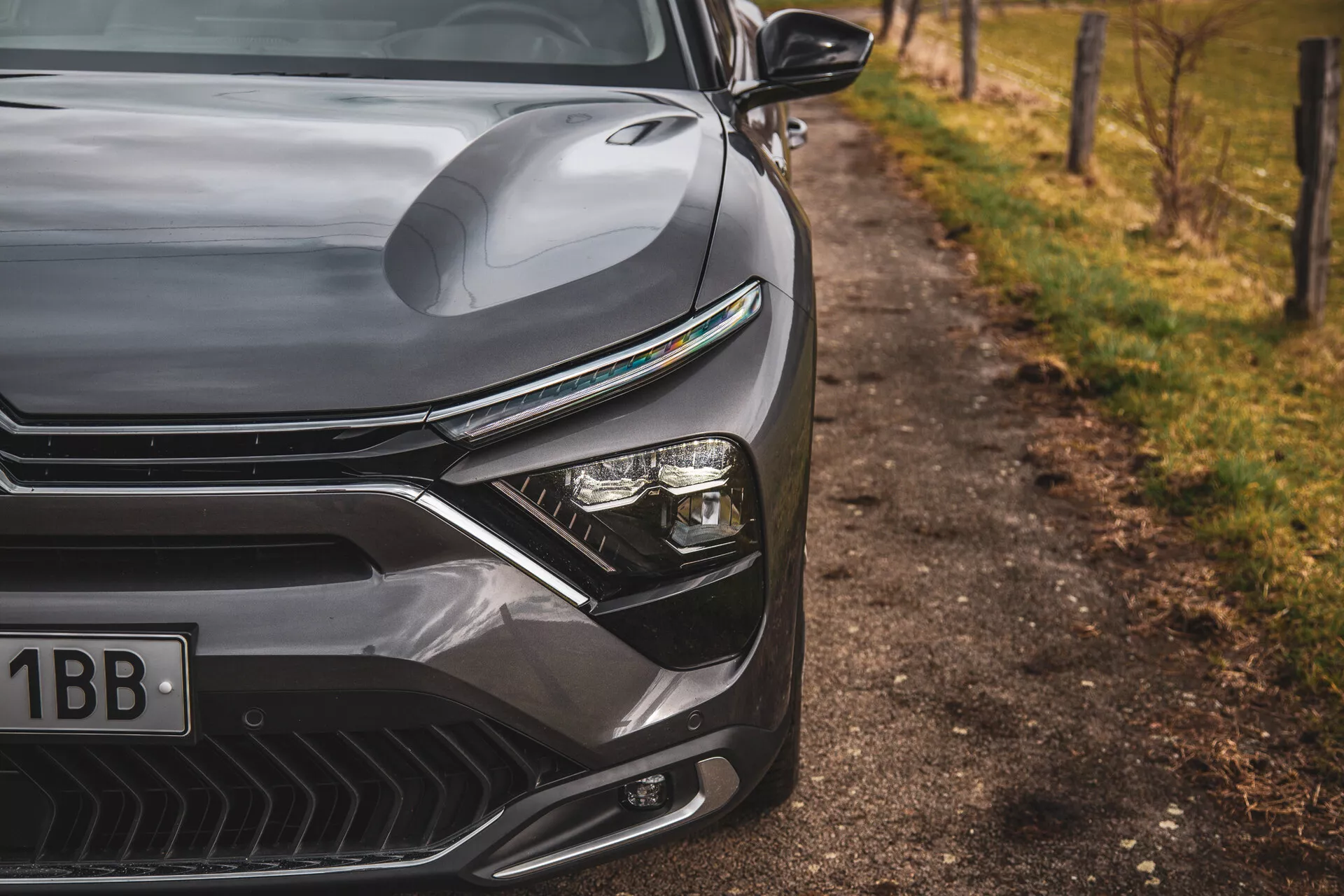 Detailný pohľad na Citroen C5-X 2022 predvádzajúci elegantný dizajn prednej ľavej strany, vrátane kontúrovanej kapoty, výraznej mriežky s horizontálnymi líniami, pokročilého systému LED svetlometov a aerodynamicky tvarovaného predného nárazníka s otvormi na prívod vzduchu, proti rozmazanému prírodnému pozadiu pri ceste.