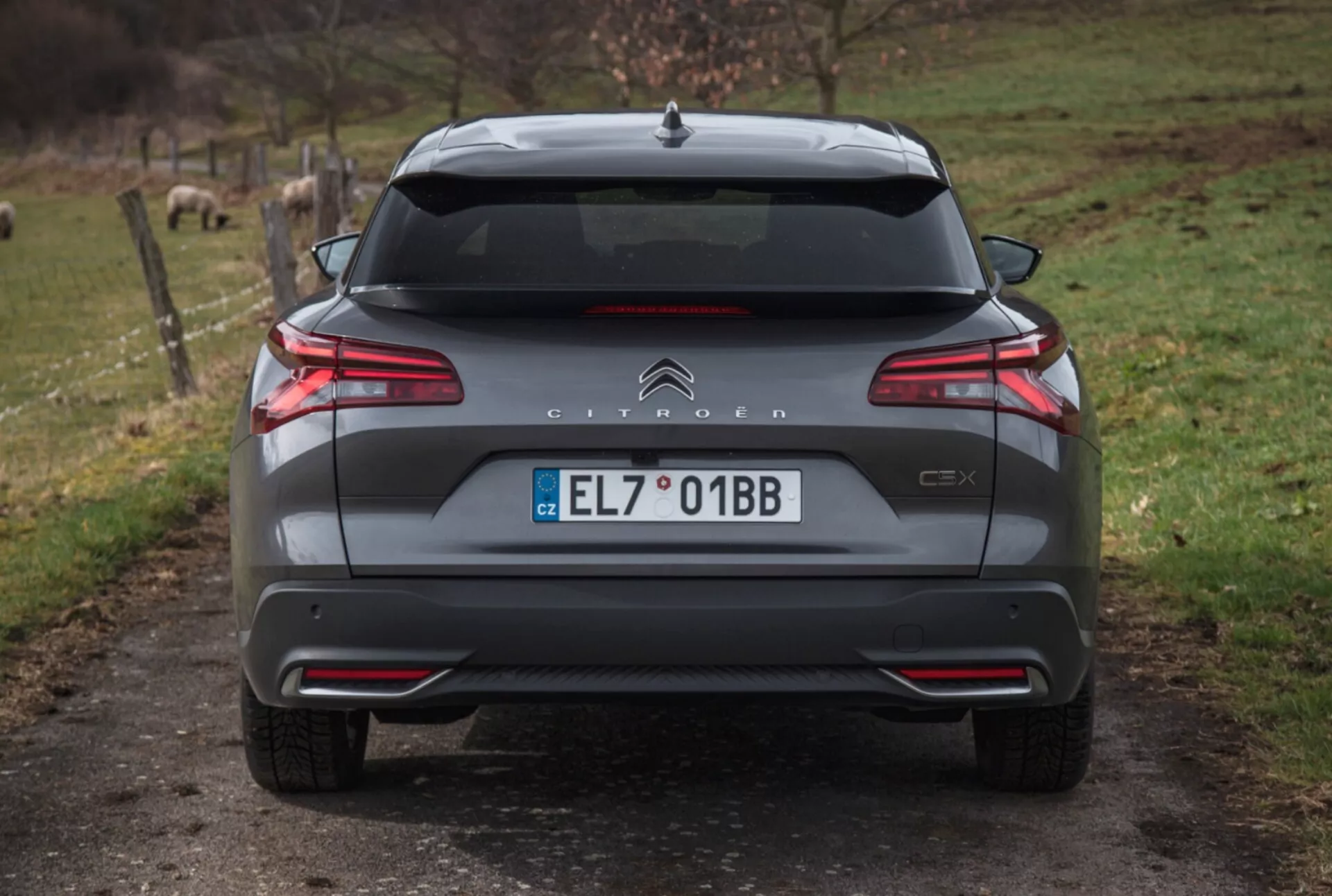 Zadný pohľad na Citroën C5X 2022 na štrkovej ceste s prírodným pozadím, predvádzajúci LED zadné svetlá, kontrolu vzdialenosti pri parkovaní, dvojitý výfuk a ďalšie vonkajšie detaily.