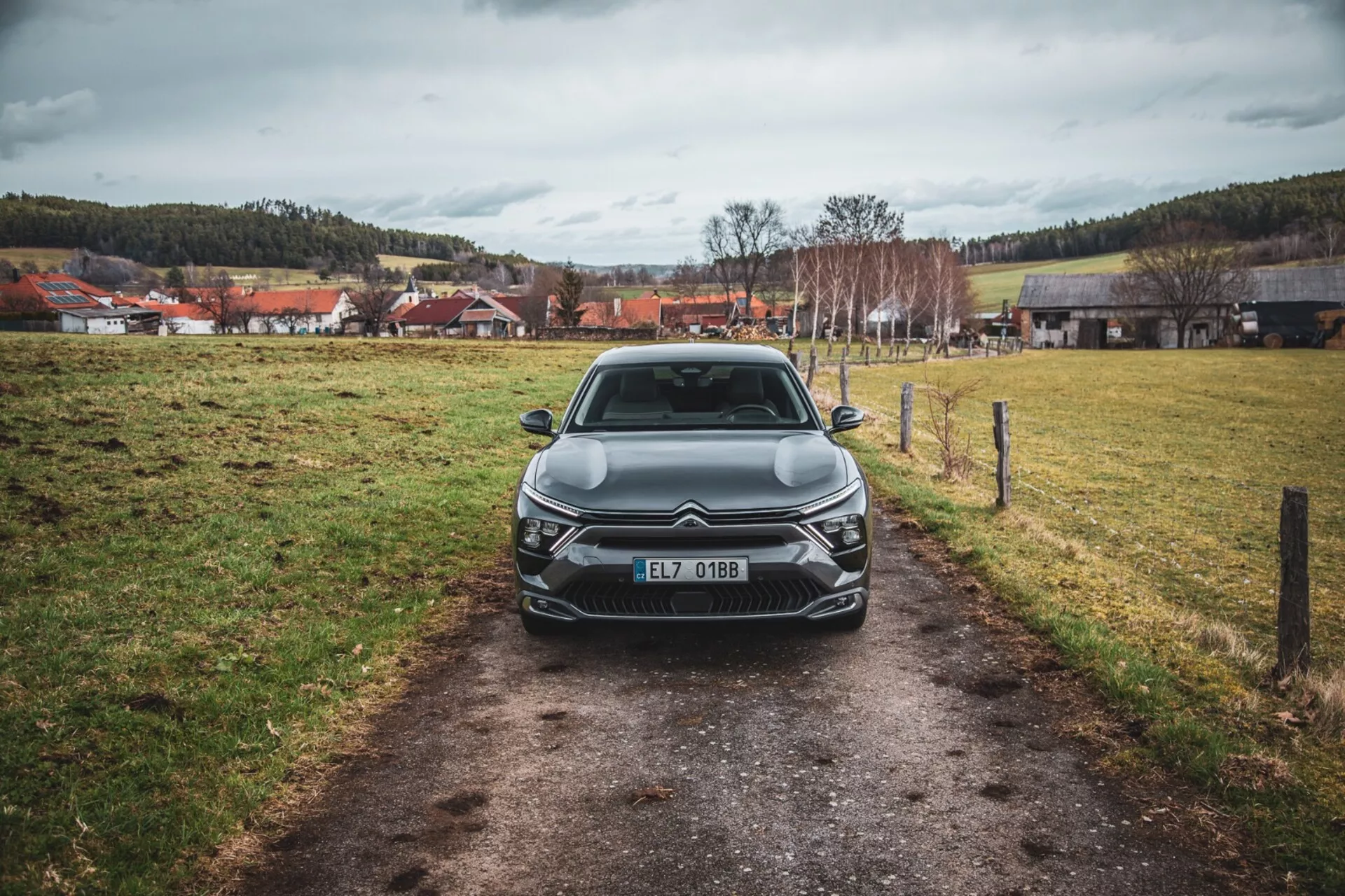 Čelný pohľad na Citroën C5-X 2022 na úzkom chodníku v trávnatom poli pod zamračenou oblohou, predvádzajúci jeho elegantný dizajn, zložitú mriežku s emblémou, uhlové LED svetlomety, sochárske línie kapoty a doplnkové predné osvetlenie, zaregistrovaný v Českej republike.