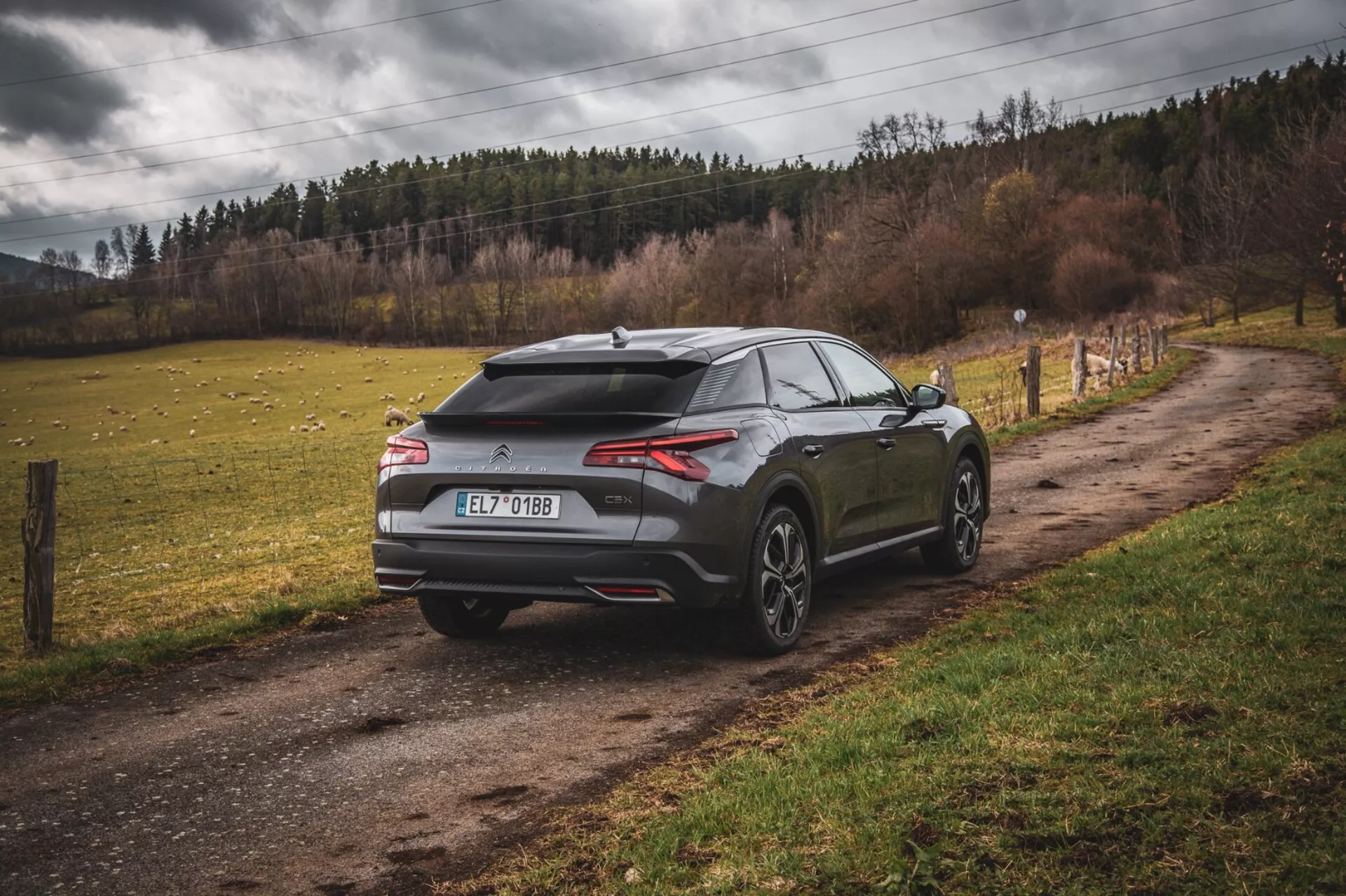 Moderné hatchback Citroen C5-X 2022 zaparkované na nezpevnenom vidieckom ceste, z pohľadu trojštvrte zozadu predvádzajúce aerodynamický dizajn so spojlerom a anténou v tvare žraločej plutvy, tenké vodorovné zadné svetlá a veľké kolesá s viacerými paprskami, naznačujúce výkon a luxus mimo mestské prostredie.