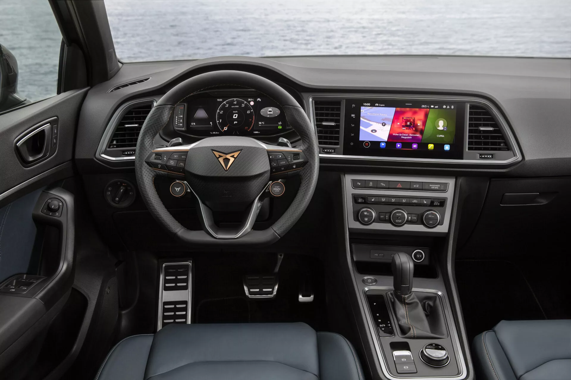 Vnútorný pohľad na Cupra Ateca 2020 zo zadného sedadla s prehľadom palubnej dosky, multifunkčného volantu s emblémom výrobcu, digitálneho prístrojového panela, dotykového displeja infotainmentu orientovaného k vodičovi, tlačidiel klimatizácie a moderného voliča prevodovky