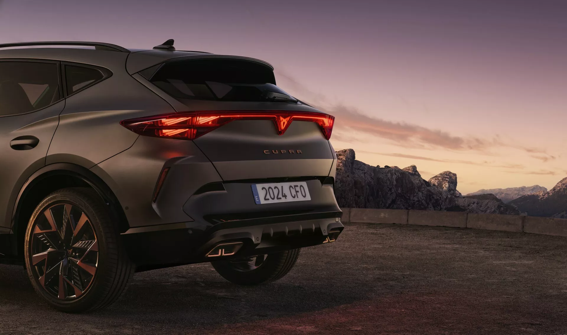 Pohľad zozadu z troch štvrtín na moderné SUV CUPRA Formentor 2024 (prvá generácia facelift) zachytený z nižšieho uhla, zvýrazňujúci jeho LED zadné svetlá, krídlový dizajn zadných svetiel a centrálne umiestnený znak CUPRA na zadných dverách. Tmavo sfarbené vozidlo, fotografované za úsvitu alebo súmraku s horským pozadím, zdôrazňuje jeho športový zadný kufor, naklonené zadné okno, viaclúčové zliatinové kolesá a vhodný dezén pneumatiky pre mestské aj mierne terénne použitie.