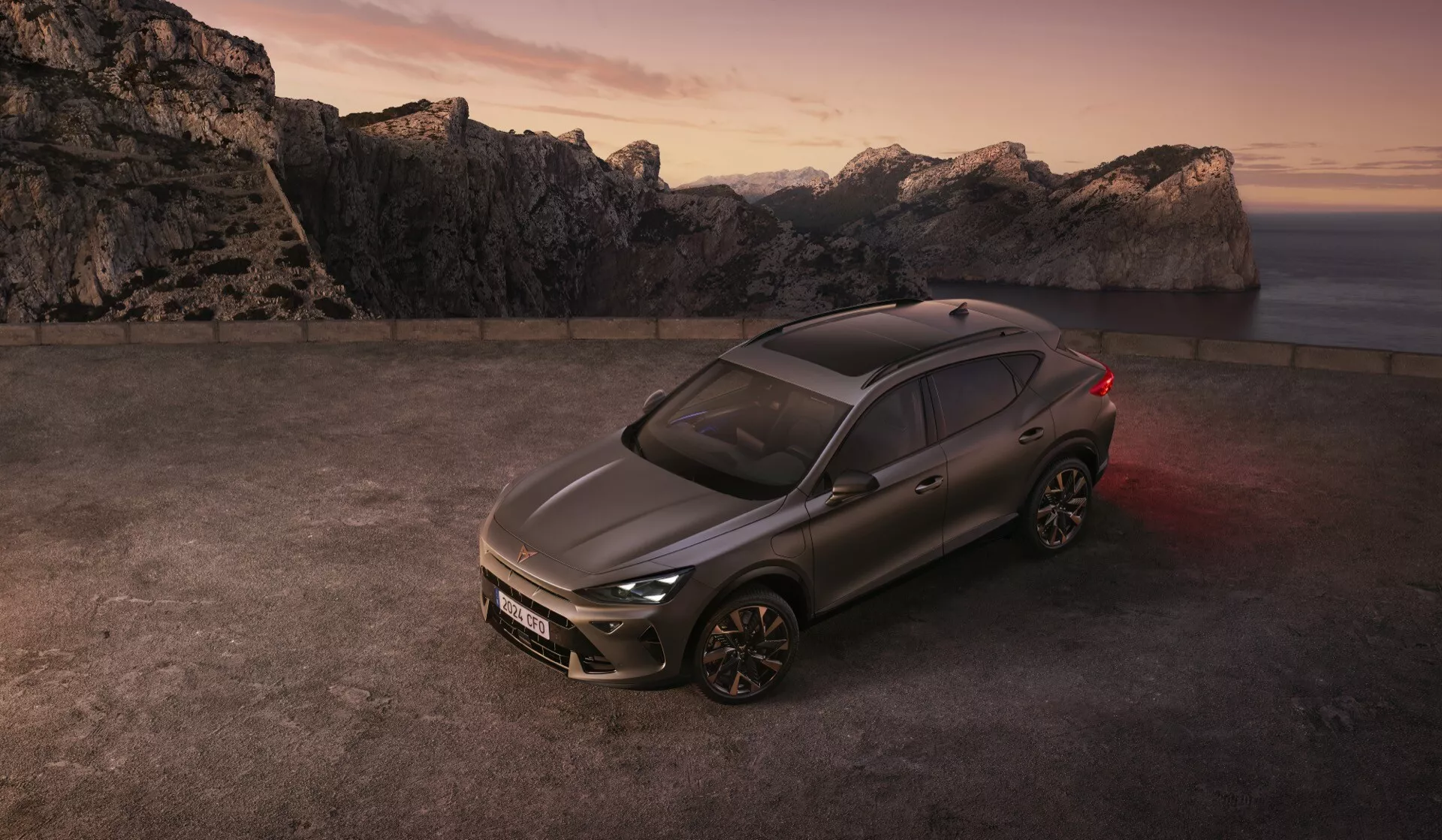 Bočný profil SUV Cupra Formentor 2024 (prvá generácia facelift) s kovovým šedým povrchom, zaparkovaný na otvorenom priestranstve s dramatickým pozadím hôr a súmraku. Zvýšený uhol ukazuje líniu strechy, trojštvrťový pohľad na predok, kolesá, dvere, prednú mriežku, LED svetlomety, zliatinové viaclúčové kolesá, čiastočne osvetlené LED zadné svetlo, tónované okná, strešné okno a kontrastnú výzdobu.