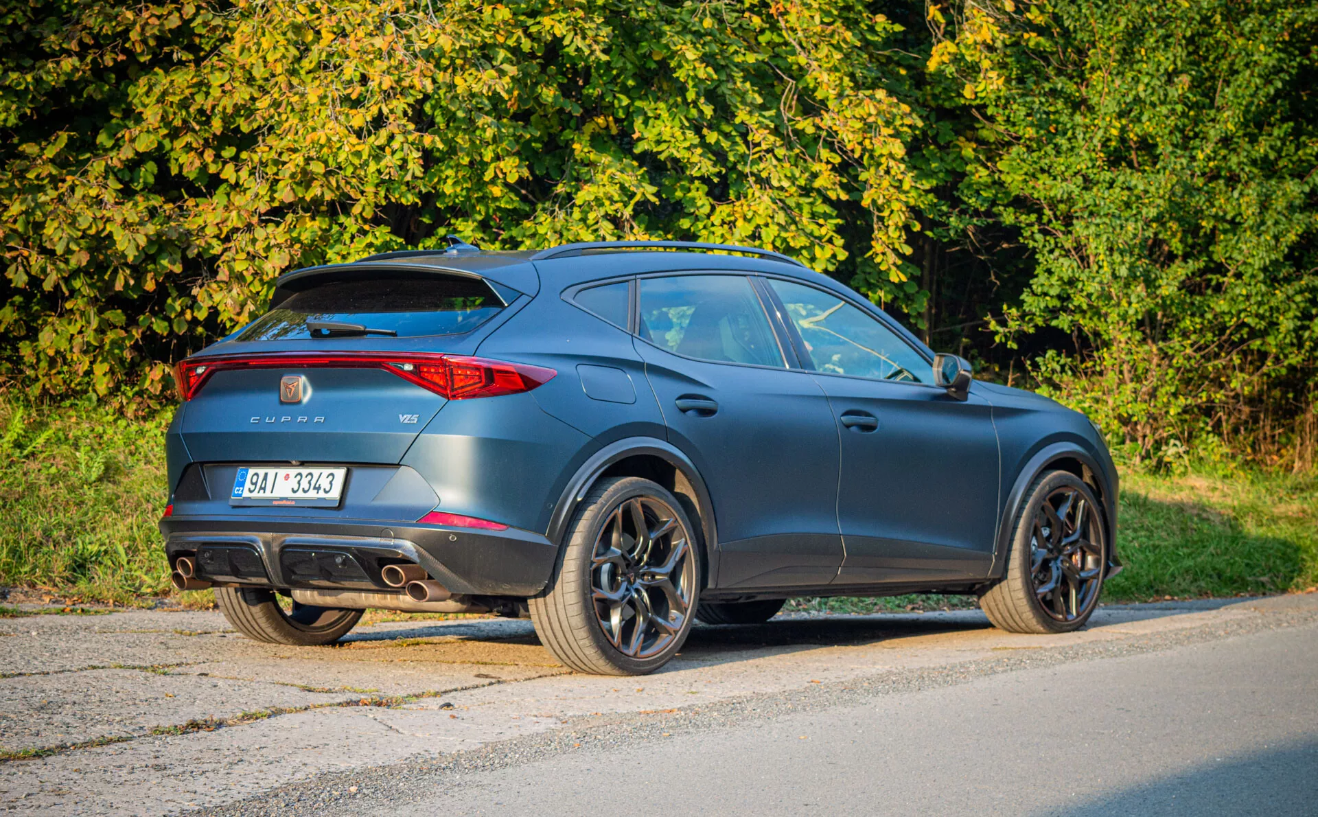 Moderné tmavošedé matné čierne crossover SUV Cupra Formentor 2020 zaparkované na asfalte, zobrazujúce zadný trojštvrťový pohľad zdôrazňujúci dizajn podobný kupé, športovú aerodynamiku, LED zadné svetlá, zadný spojler, dvojité výfuky a veľké zliatinové kolesá v predmestskom prostredí