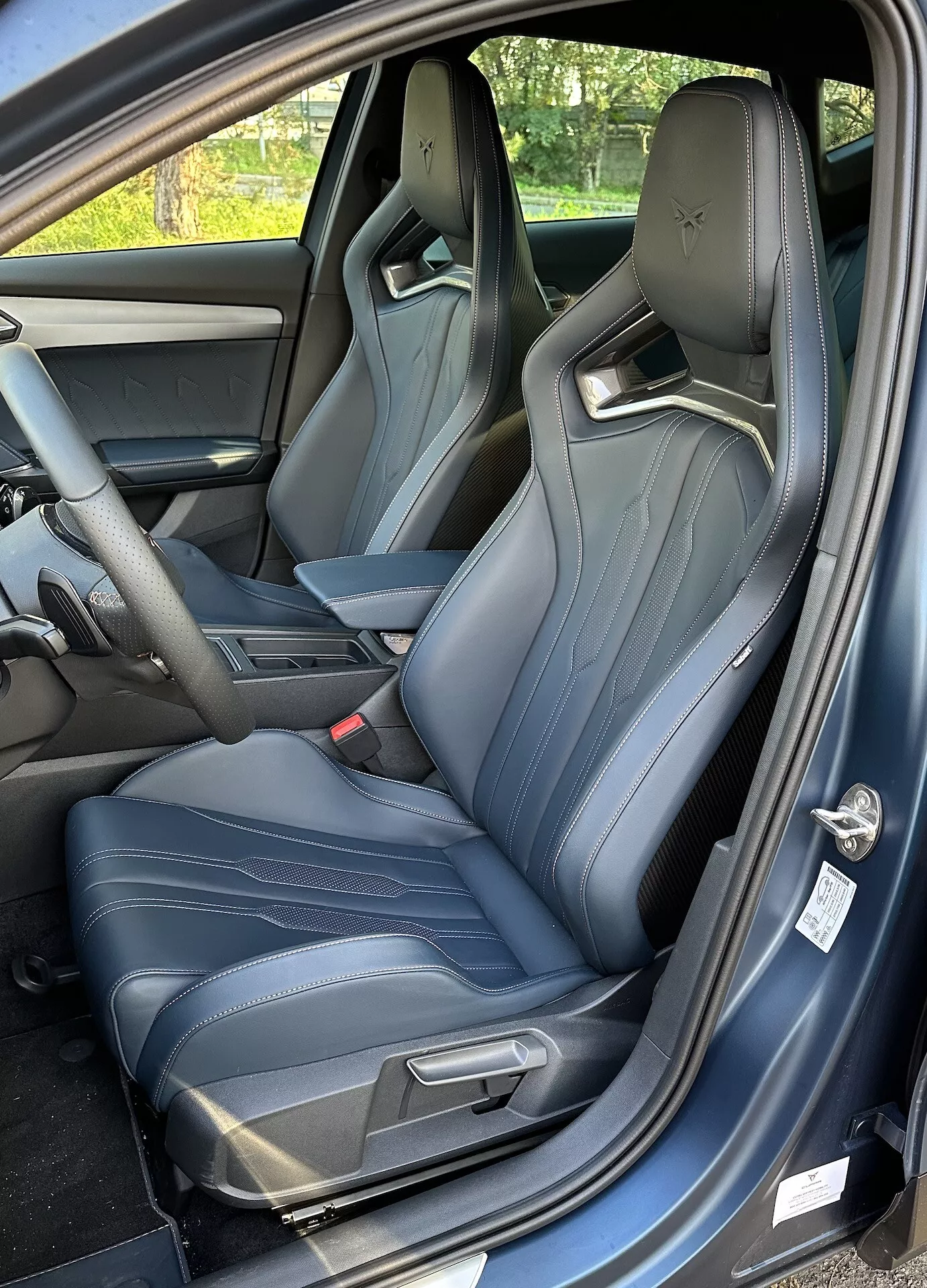 Pohľad do interiéru Cupra Formentor 2020, zobrazuje sedadlo vodiča z perspektívy troch štvrtín, s otvorenými dverami, športovými sedadlami s ľahkým modrým šitím a náhľadom na centrálnu konzolu a palubnú dosku.