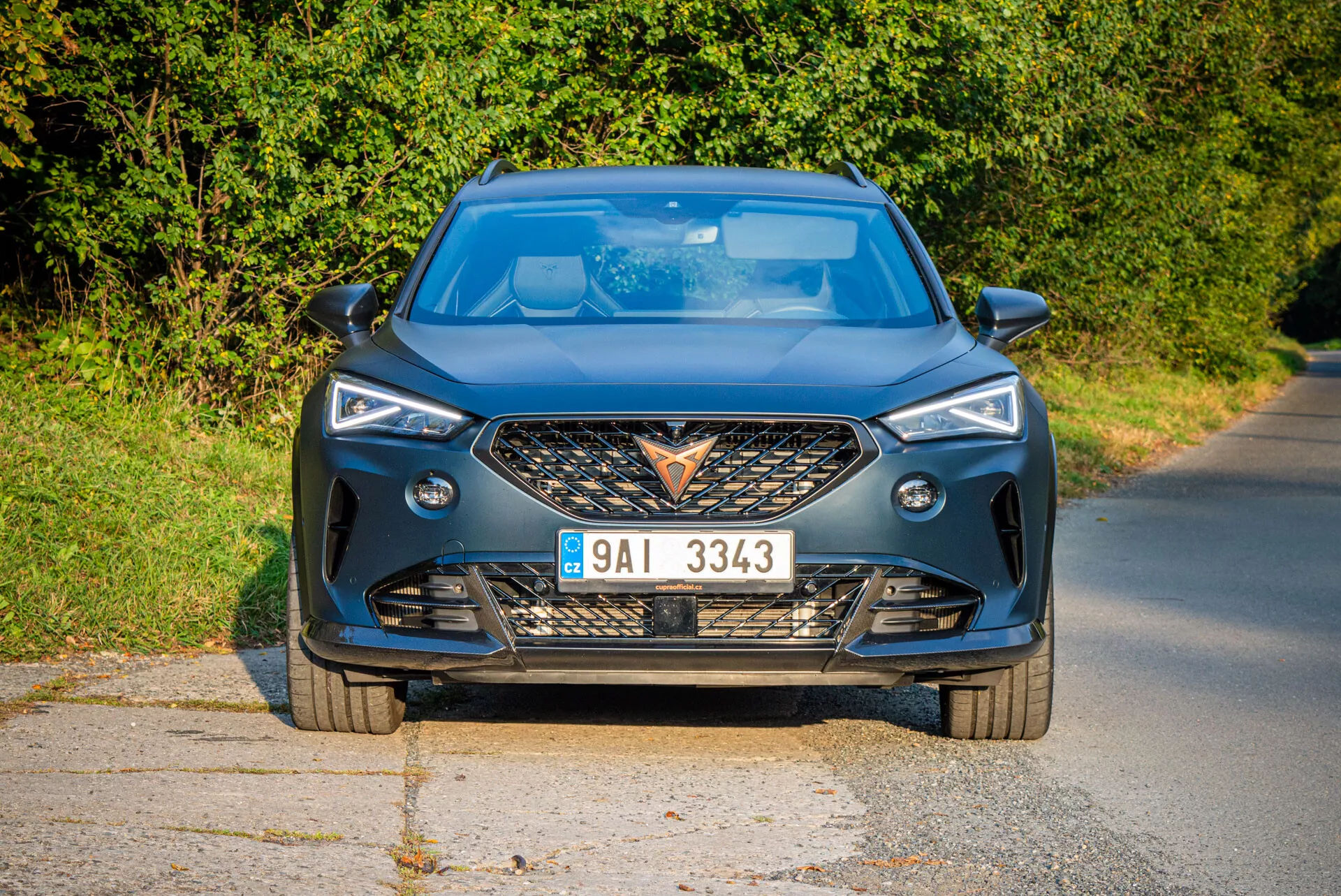 Moderný pohľad z prednej strany na Cupra Formentor 2020 na spevnenej ceste s prírodným denným svetlom a vidieckym pozadím, zdôrazňujúci agresívnu mriežku chladiča, ostré LED svetlomety, dynamické kontúry kapoty a pokročilé technologické funkcie