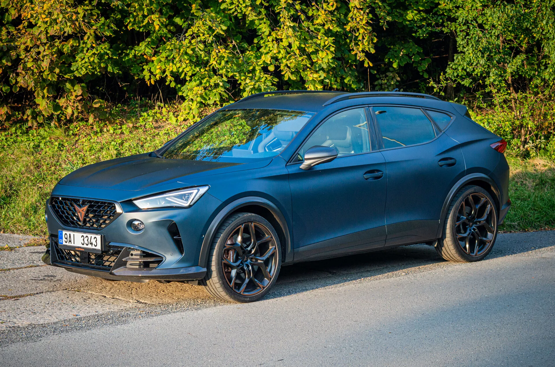 Modré crossover SUV Cupra Formentor 2020 zaparkované pri ceste s vegetáciou na pozadí, zvýrazňujúce pohľad z tretej štvrtiny zameraný na stranu vodiča, prednú mriežku, uhlové svetlomety, viacramenné zliatinové kolesá a športový bočný profil ako kupé