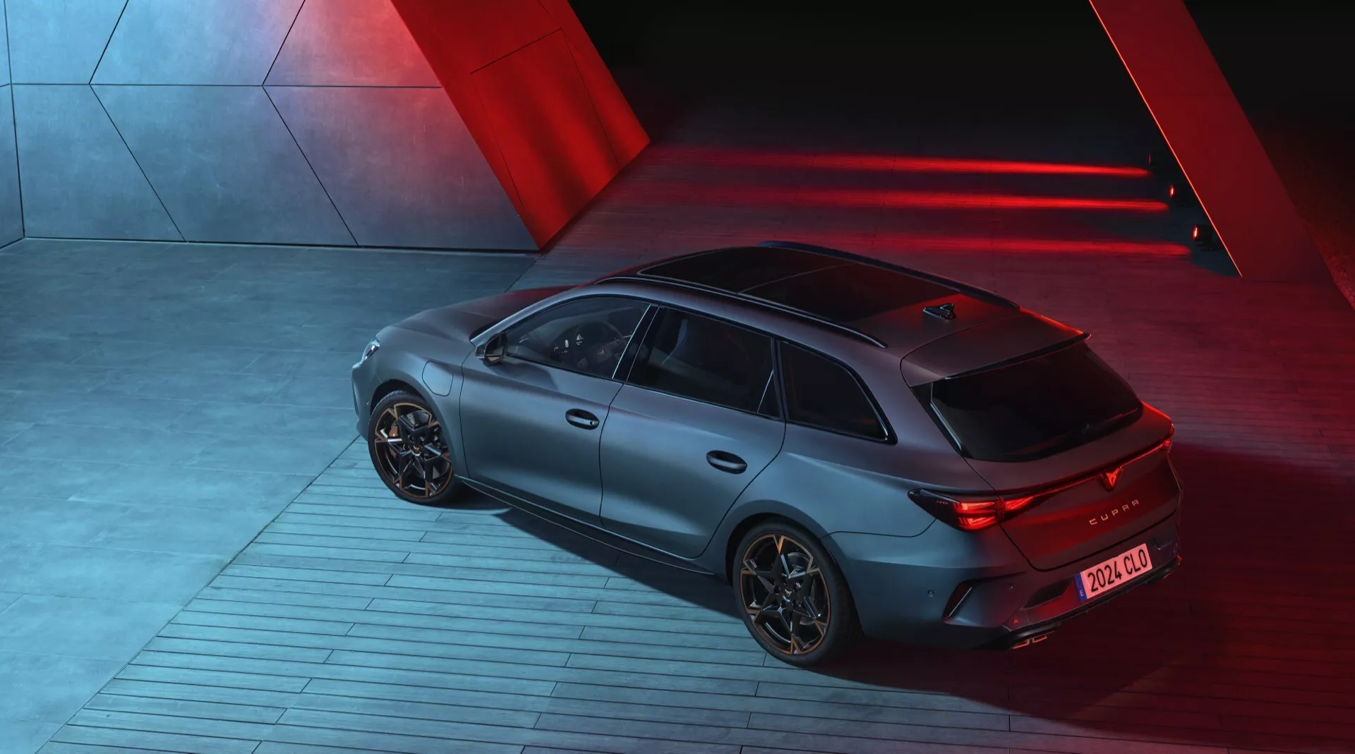 Pohľad z výšky na model Cupra Leon 2024 (1. generácia facelift) nasnímaný v noci, zobrazujúci zadnú a pravú stranu športového hatchbacku. Auto je osvetlené dramatickými červenými smerovými svetlami, ktoré vytvárajú elegantnú, futuristickú atmosféru. Má matný povrch, osvetlené LED zadné svetlá a aerodynamické hliníkové kolesá. Vozidlo stojí na vzorovanej priemyselnej ploche, naznačujúcej pohyb. Viditeľné technologické prvky zahŕňajú zadný difuzér a krátku anténu smerom k zadnej časti pre komunikačné technológie.