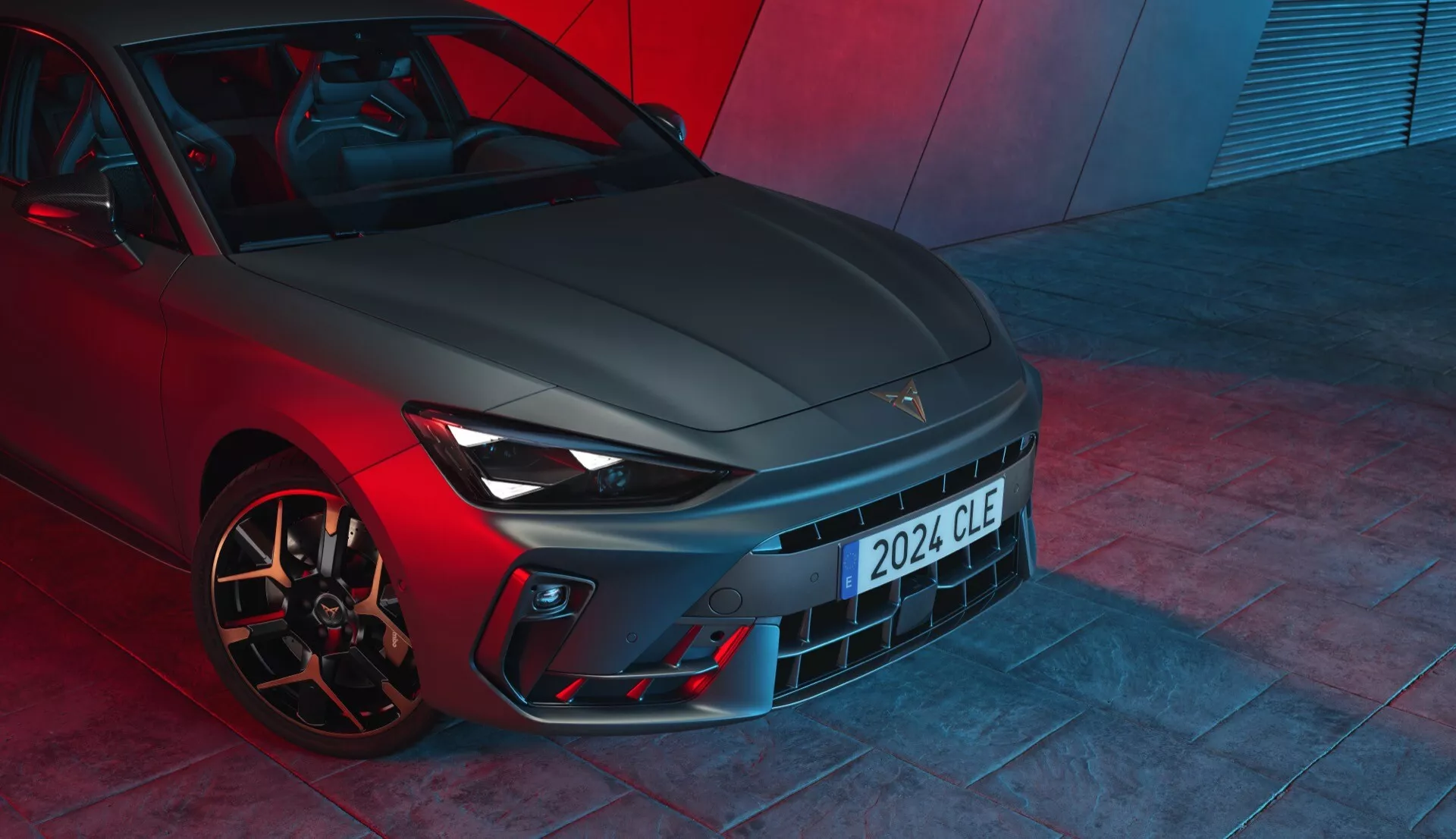 Pohľad z nízkeho uhla na prednú časť vozidla Cupra Leon 2024 (facelift 1. generácie), predvádzajúci jeho dizajnové prvky s výraznou maskou, elegantnými svetlometmi s LED denným osvetlením, športovým nárazníkom s vetracími otvormi a výkonovými zliatinovými kolesami s vysokovýkonnými brzdovými strmeňmi. Kovový lak a dramatické červené osvetlenie vytvárajú dojem vysokovýkonného vozidla, pravdepodobne fotografovaný na podlahe showroomu.