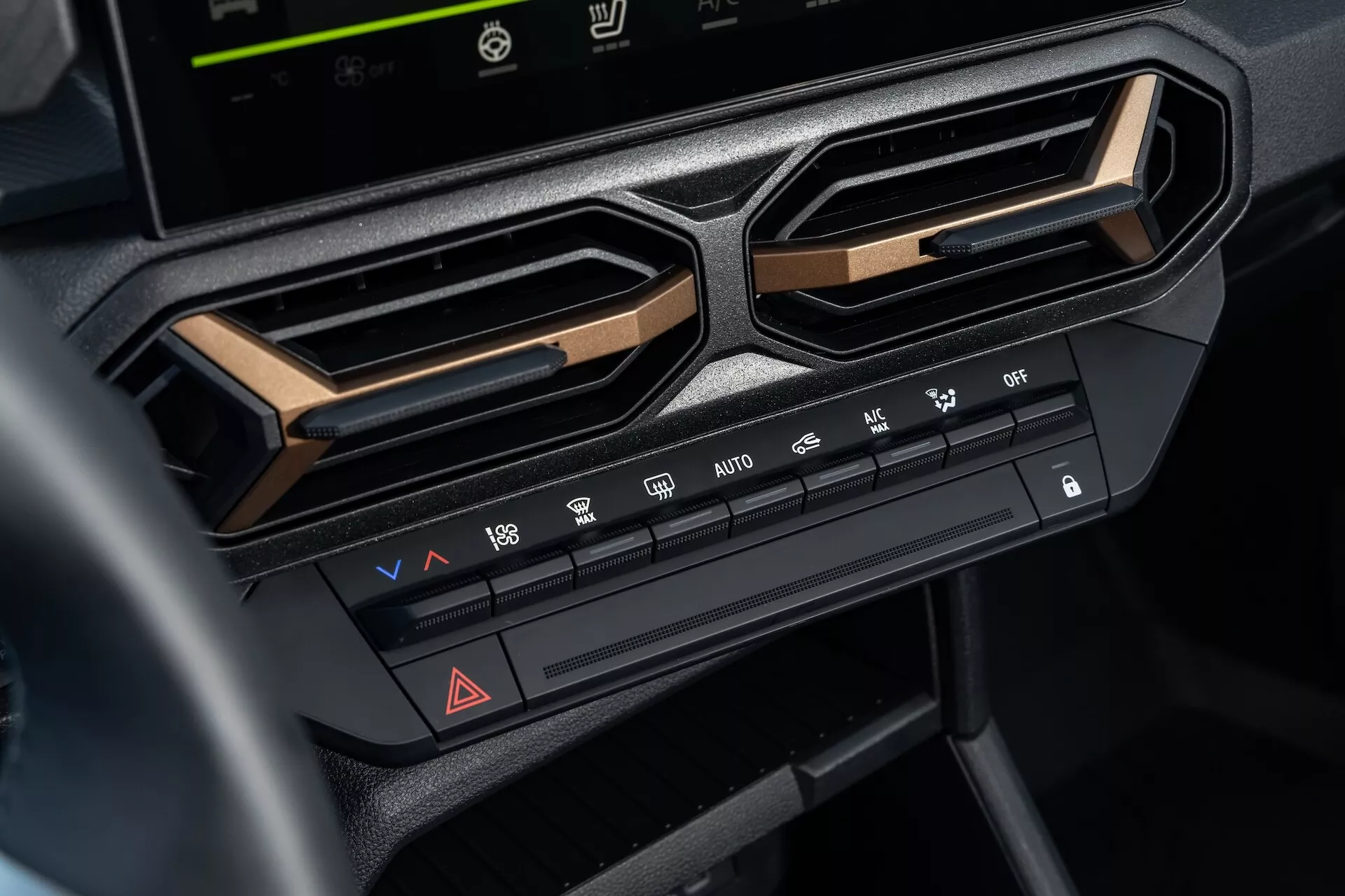 Detailný pohľad na systém klímy v Dacia Duster 3. generácie 2024 na stredovej konzole pod obrazovkou infotainmentu, zahŕňa ovládanie teploty a rýchlosti ventilátora, funkcie distribúcie vzduchu, odmrazovacie funkcie, tlačidlo výstražných svetiel a možnosti klimatizácie, orámované uhlovými ventilačnými mriežkami a zdôraznené kovovým lemom pre moderný estetický vzhľad interiéru