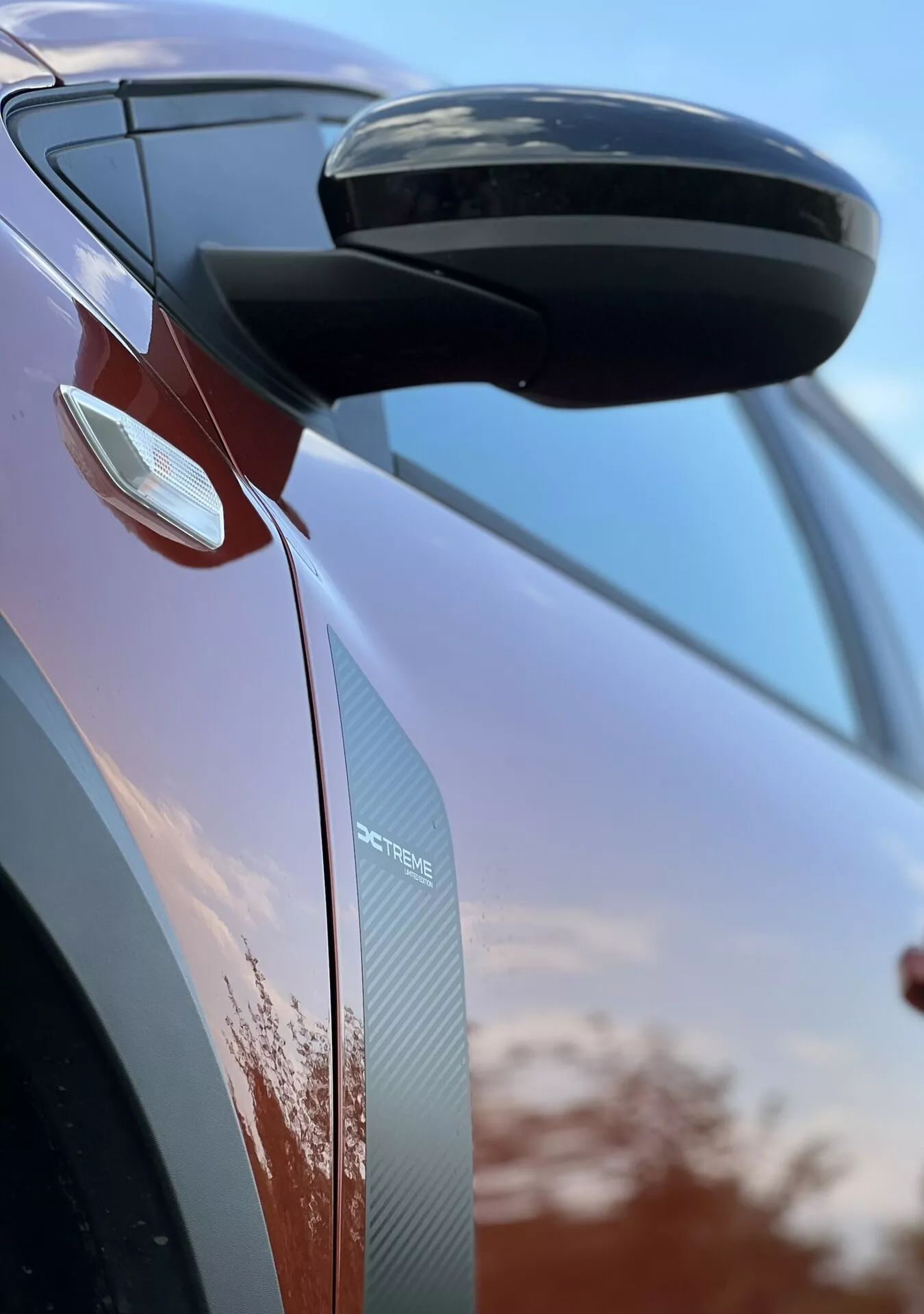 Detailný pohľad na Dacia Jogger 2022 zameraný na bočné spätné zrkadlo a dizajn dverí, ktorý predvádza aerodynamické prvky, integrované smerové svetlá a textúry z uhlíkových vlákien na pozadí oblohy