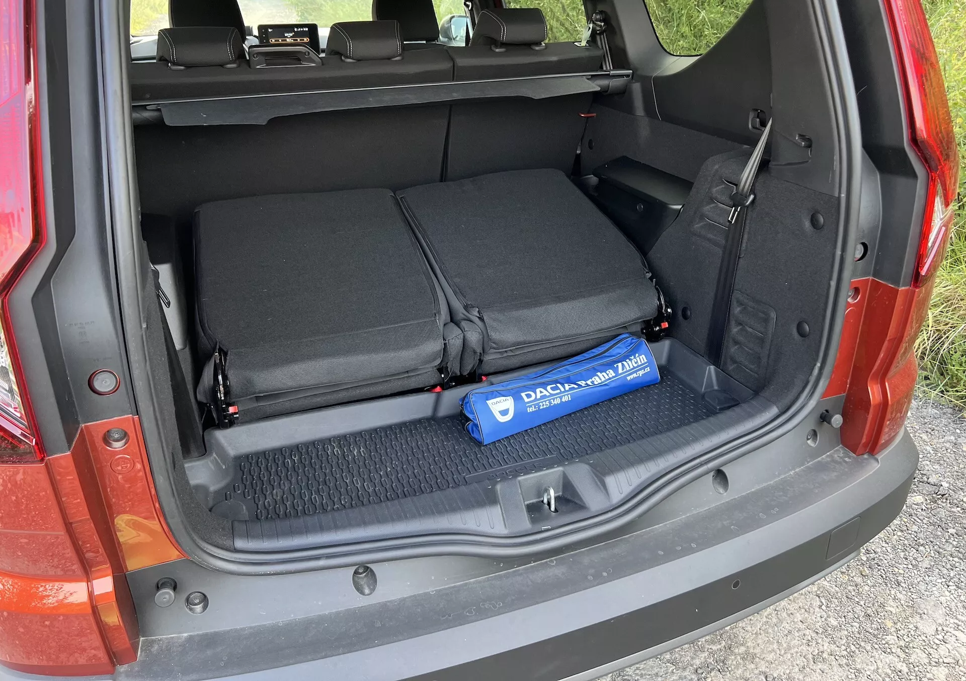 Detail batožinového priestoru Dacia Jogger 2022 s otvoreným nákladovým priestorom, sklopenými zadnými sedadlami, textilnými slučkami na zabezpečenie predmetov a modrým valcom Decaux na kobercovanej podlahe