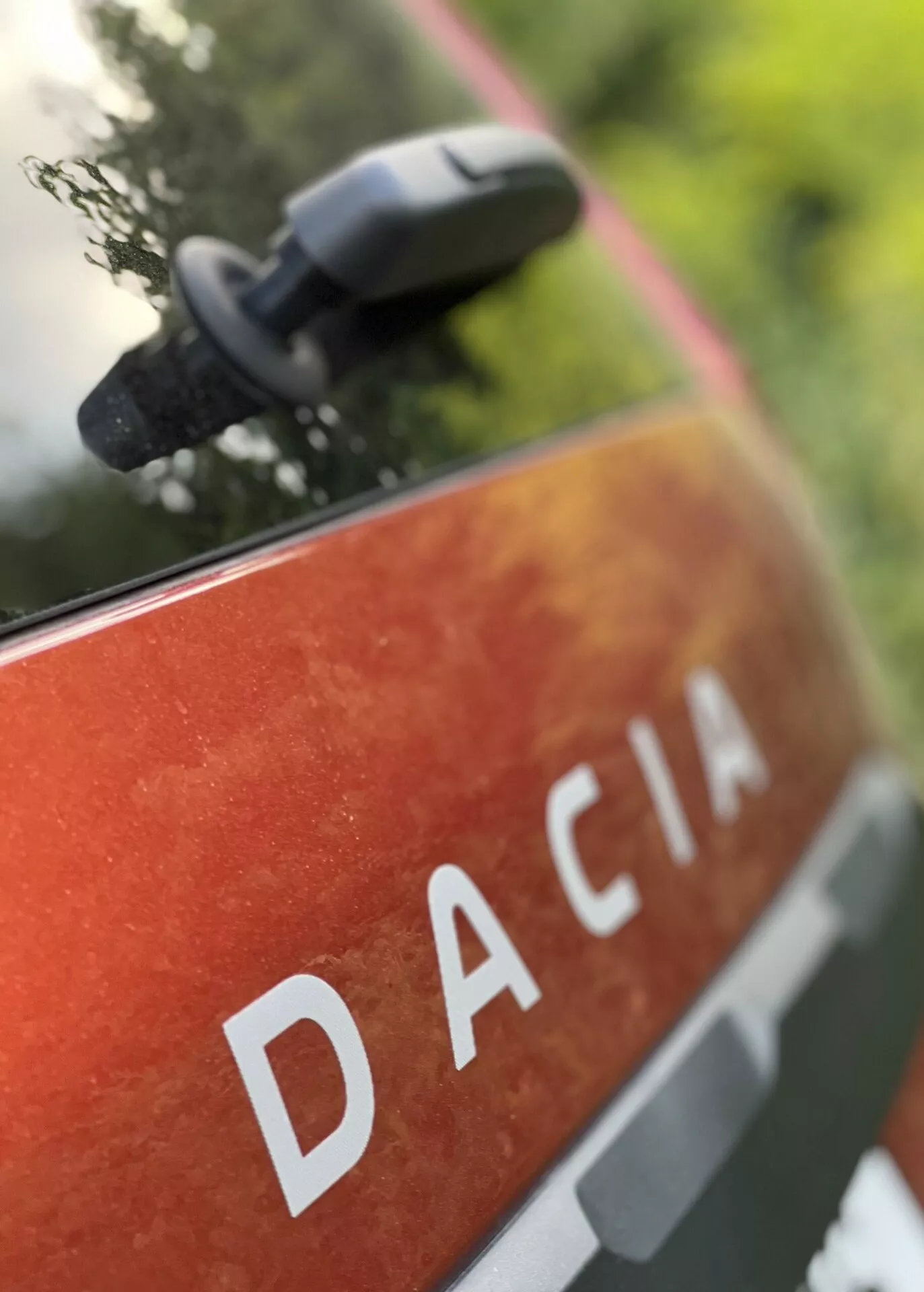 Detailný pohľad na zadný panel Dacia Jogger 2022 s nápisom DACIA veľkými bielymi písmenami na metalickej oranžovo-červenej farbe s bodkami a nečistotami, naznačujúci vonkajšie použitie a odolnosť, umiestnený v prírodnom prostredí s rozmazaným bočným zrkadlom naznačujúcim moderné funkcie zrkadla.