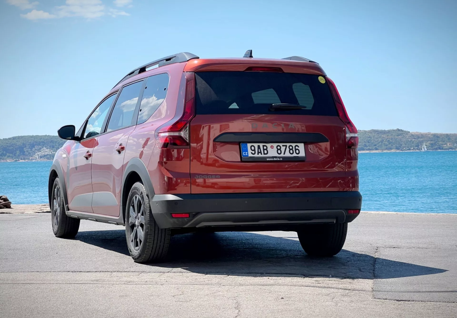 Dacia Jogger 2022 v červenej farbe zachytená z trojštvrťového zadného uhla, ktorý ukazuje bok a dizajn zadných dverí SUV, zaparkované pri vodnej ploche s horizontom na pozadí, s dôrazom na jej kolesá z ľahkých zliatin s viacerými ramenami, LED zadné svetlá a aerodynamický zadný spojler