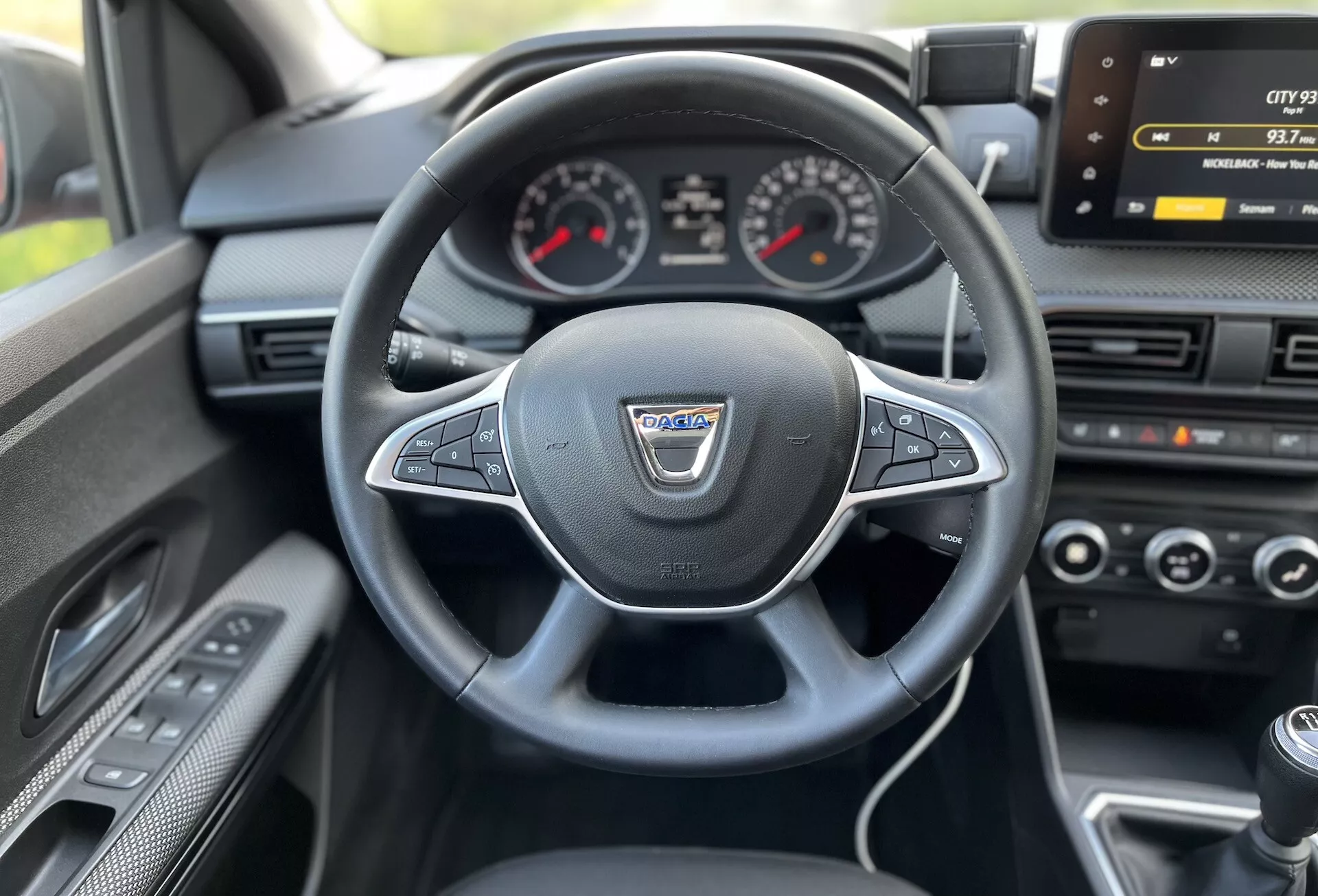 Interiérový pohľad z miesta vodiča na Daciu Jogger 2022 ukazujúci viacfunkčný volant obalený kožou, analógový prístrojový panel s centrálnym digitálnym displejom, obrazovku infotainmentu, ovládanie klimatizácie a páku manuálnej prevodovky