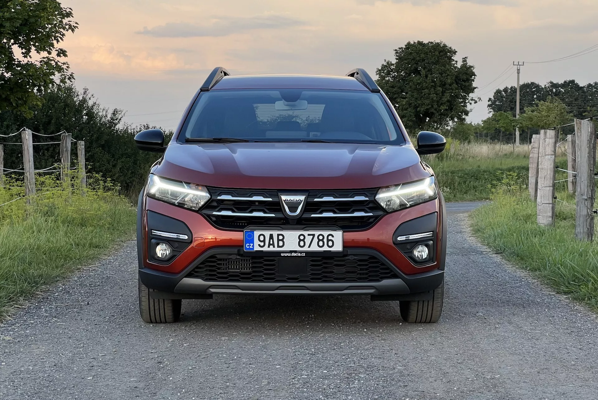 Dacia Jogger 2022 umiestnená na úzkej vidieckej ceste, ponúka symetrický pohľad spredu, ktorý zvýrazňuje moderný dizajn vozidla vrátane výraznej prednej mriežky s veľkým emblémom, štíhlych LED svetlometov, konturovaných liniek kapoty, predného nárazníka s hmlovými svetlami a parkovacími senzormi, a bočných zrkadiel s integrovanými smerovými svetlami.