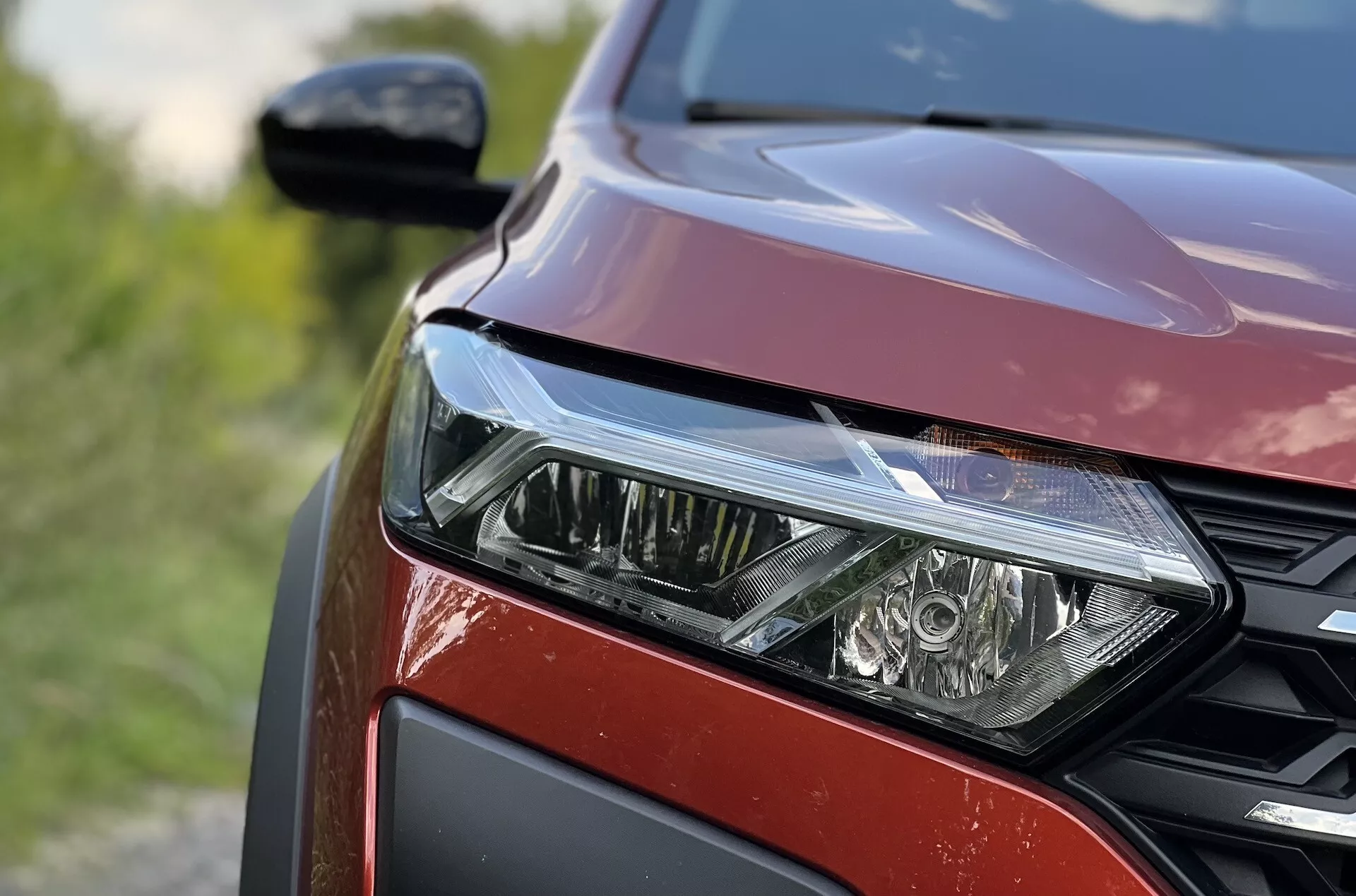 Detailný pohľad na prednú ľavú stranu Dacia Jogger 2022 zdôrazňujúci súbor LED svetlometov, vzor mriežky s medovými bunkami a metalický červený povrch laku