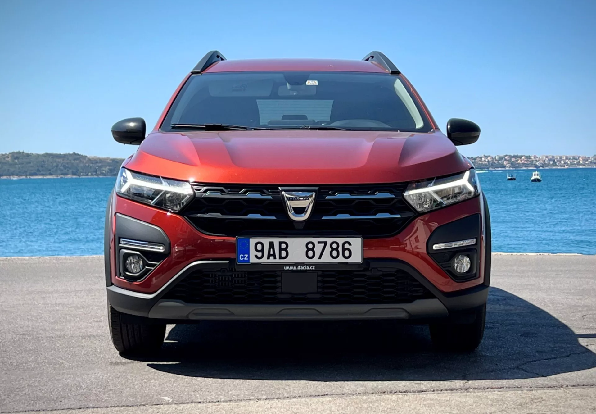 Dacia Jogger 2022 prezentuje svoj moderný dizajn crossover a pokročilé technologické funkcie v pokojnom vodnom prostredí, s výraznou mriežkou s logom, LED svetlometmi, hmlistými svetlami, pravdepodobne senzormi ADAS, vzduchovými vstupmi pre chladenie a radar, dvojfarebnou červeno-čiernou farebnou schémou, aerodynamickým kapotom, vybaveným stieračmi naznačujúcimi možnú technológiu citlivú na dažď, bočnými zrkadlami s integrovanými smerovkami pravdepodobne obsahujúcimi technológiu monitorovania mŕtveho uhla, pod jasným nebom.