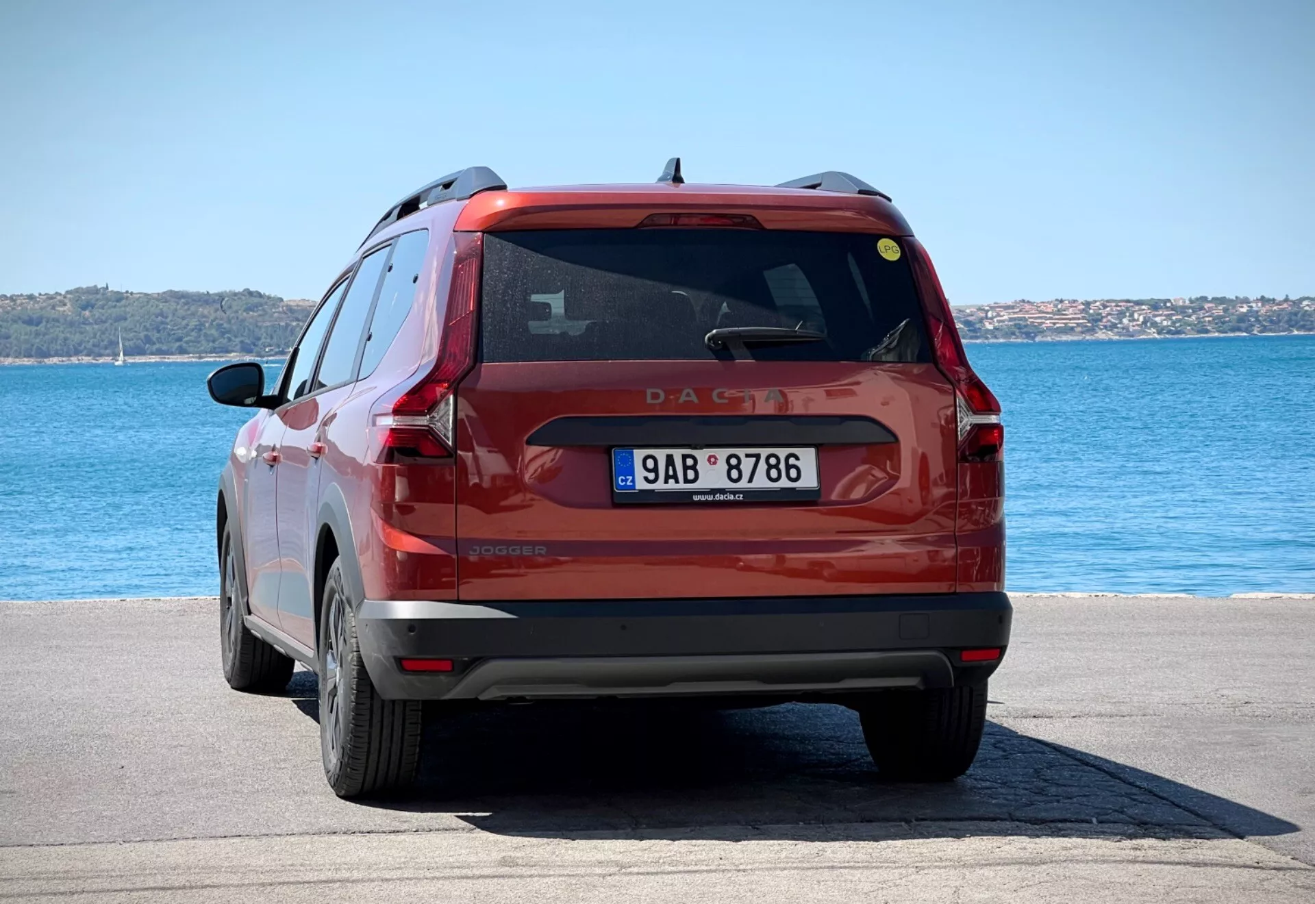 Zadný pohľad na červené kompaktné SUV Dacia Jogger 2022 zaparkované na dlaždenom nábreží pod jasným denným svetlom, predvádzajúce celú zadnú časť vozidla vrátane zadných svetiel, zadného okna a evidenčnej značky na pozadí modrej vody a jasnej oblohy.