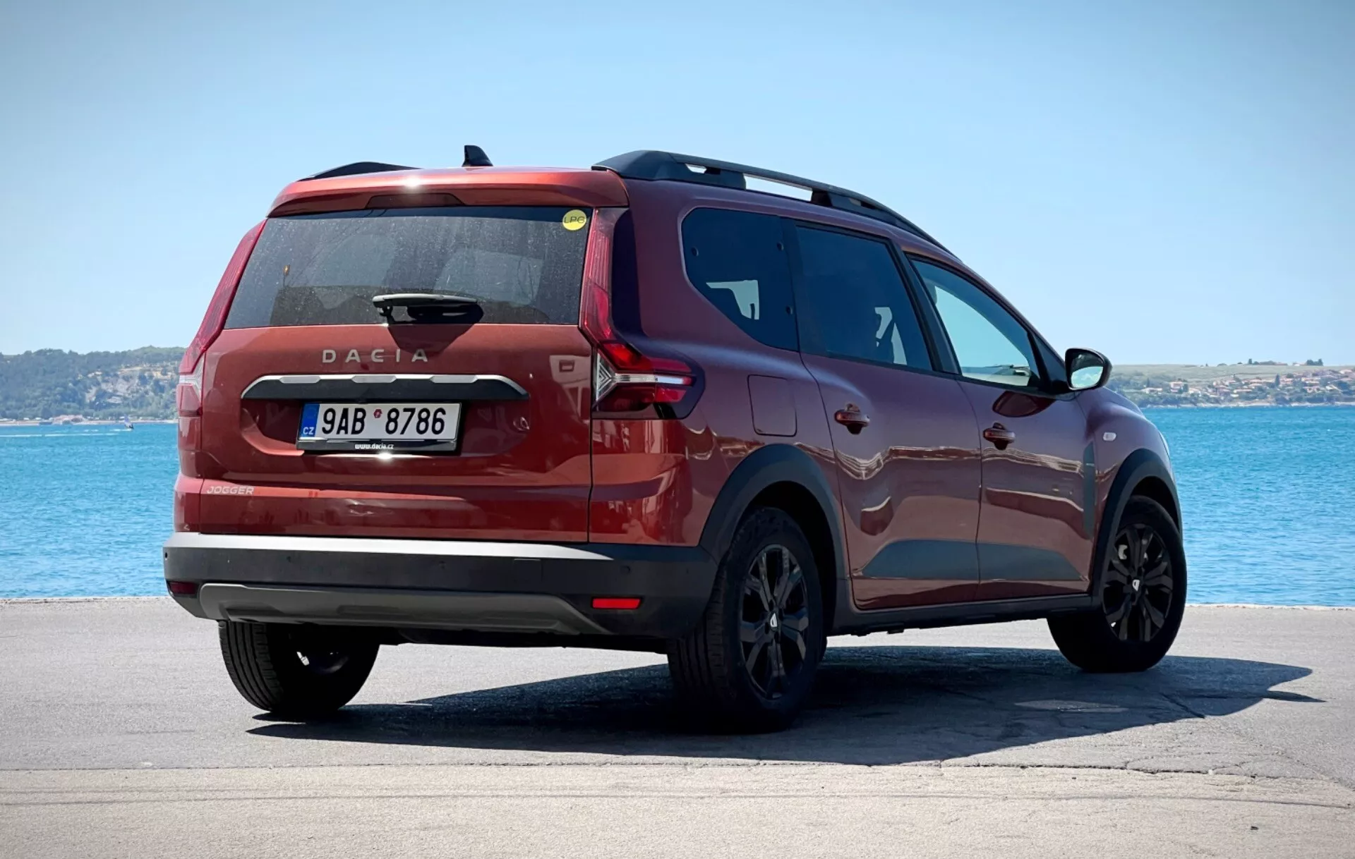 Dacia Jogger 2022 fotografovaná z trojštvrťového zadného pohľadu umiestnená na vydláždenej ploche s telom vody a jasným nebom na pozadí, zobrazuje moderné zadné svetlá, centrálne umiestnený symbol Dacie a elegantný bočný profil proti pokojnému pobrežnému alebo jazernému prostrediu.