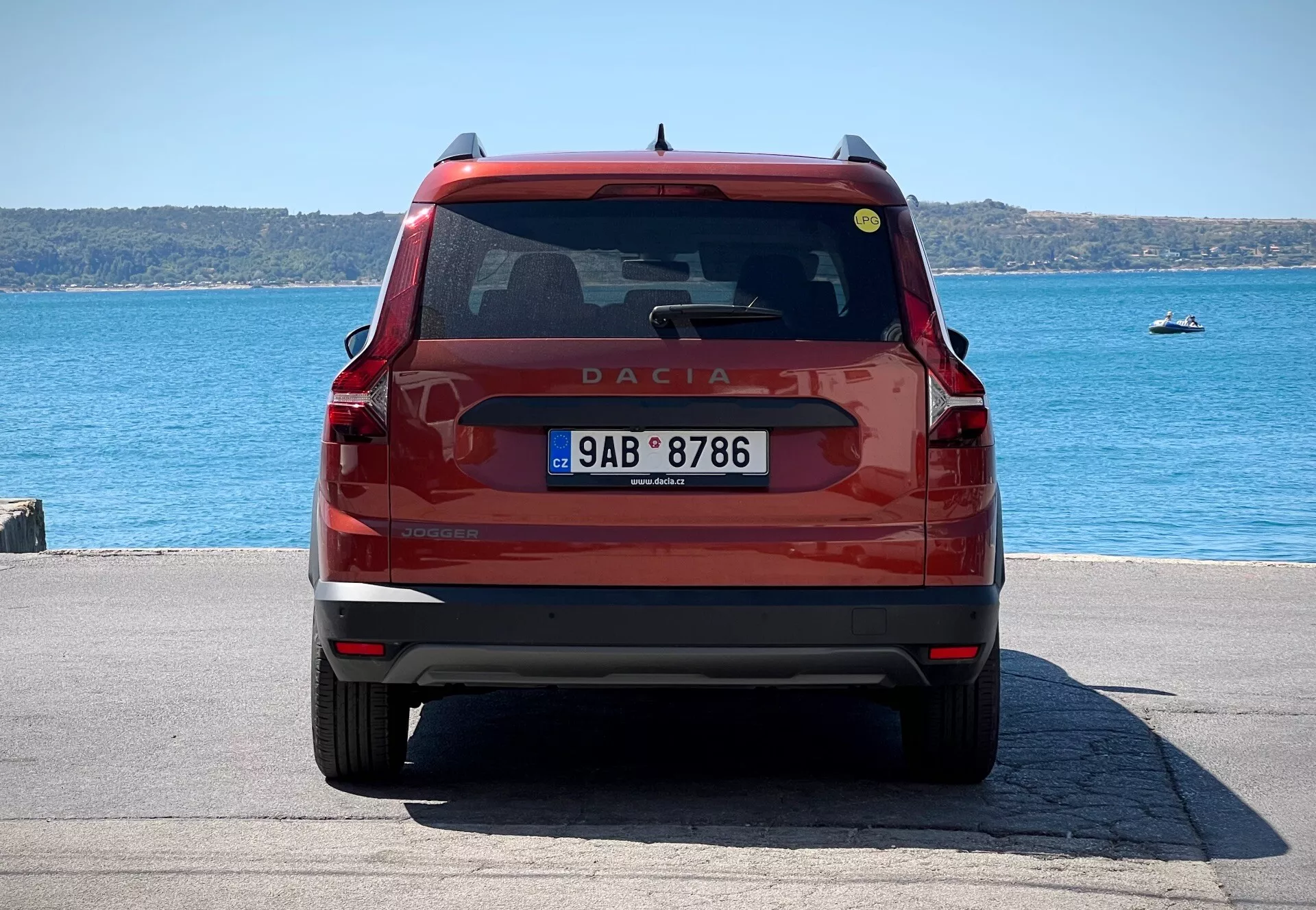 Zadný pohľad na červený Dacia Jogger 2022, ukazujúci veko batožinového priestoru s emblémom a názvom modelu, zadné čelné sklo s stieračom a brzdovým svetlom, moderné zadné svetlá, evidenčnú značku a nárazník s odrazkami a potenciálnymi parkovacími senzormi, zaparkovaný pri nábreží vo dne.