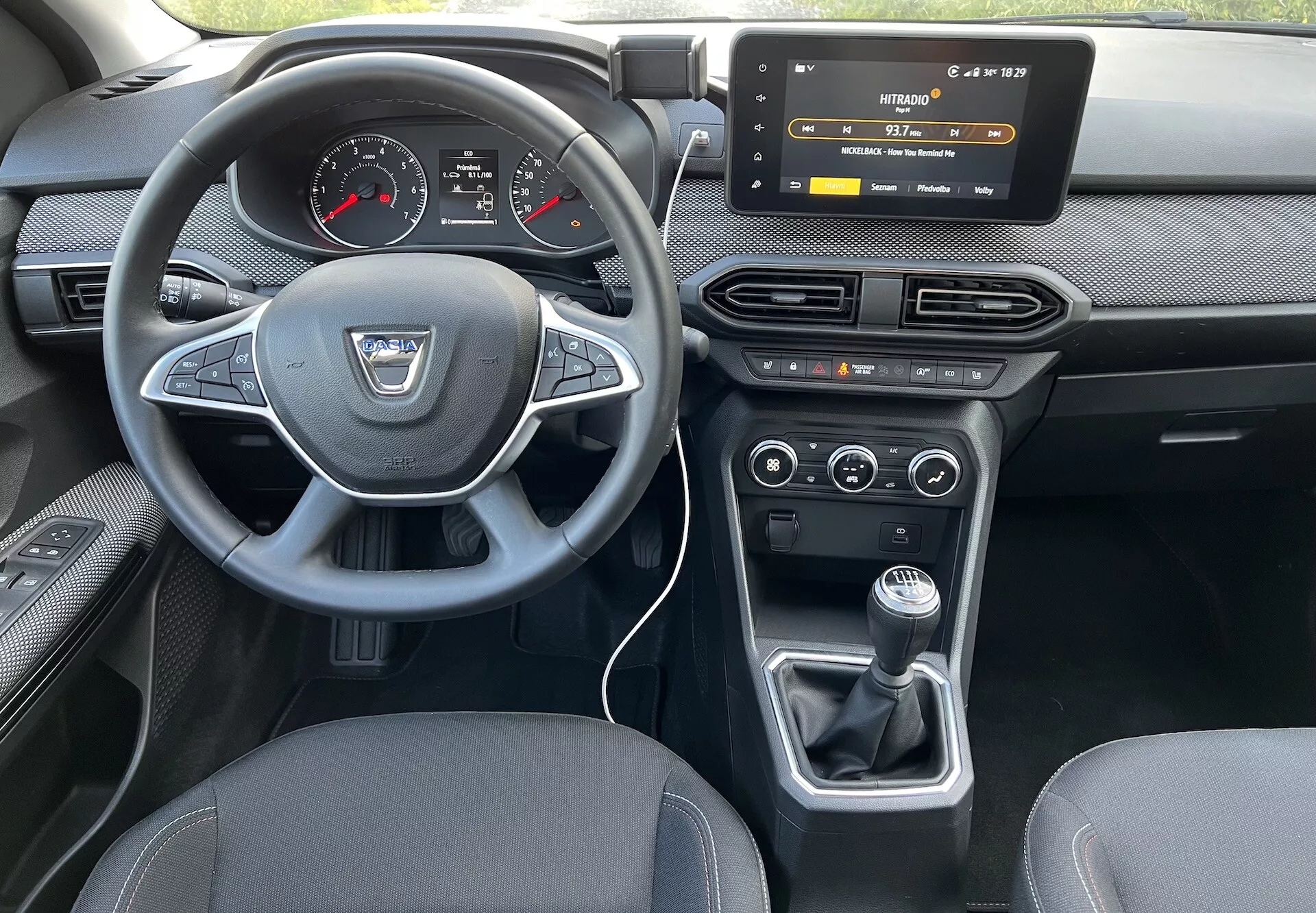 Pohľad na interiér Dacia Jogger 2022 zo zadného sedadla, zvýrazňujúci prístrojovú dosku vodiča, volant s logom výrobcu a ovládacími tlačidlami, digitálno-analógový prístrojový panel, dotykový informačný systém, ovládacie prvky HVAC, duálnu klimatizáciu a manuálnu prevodovku