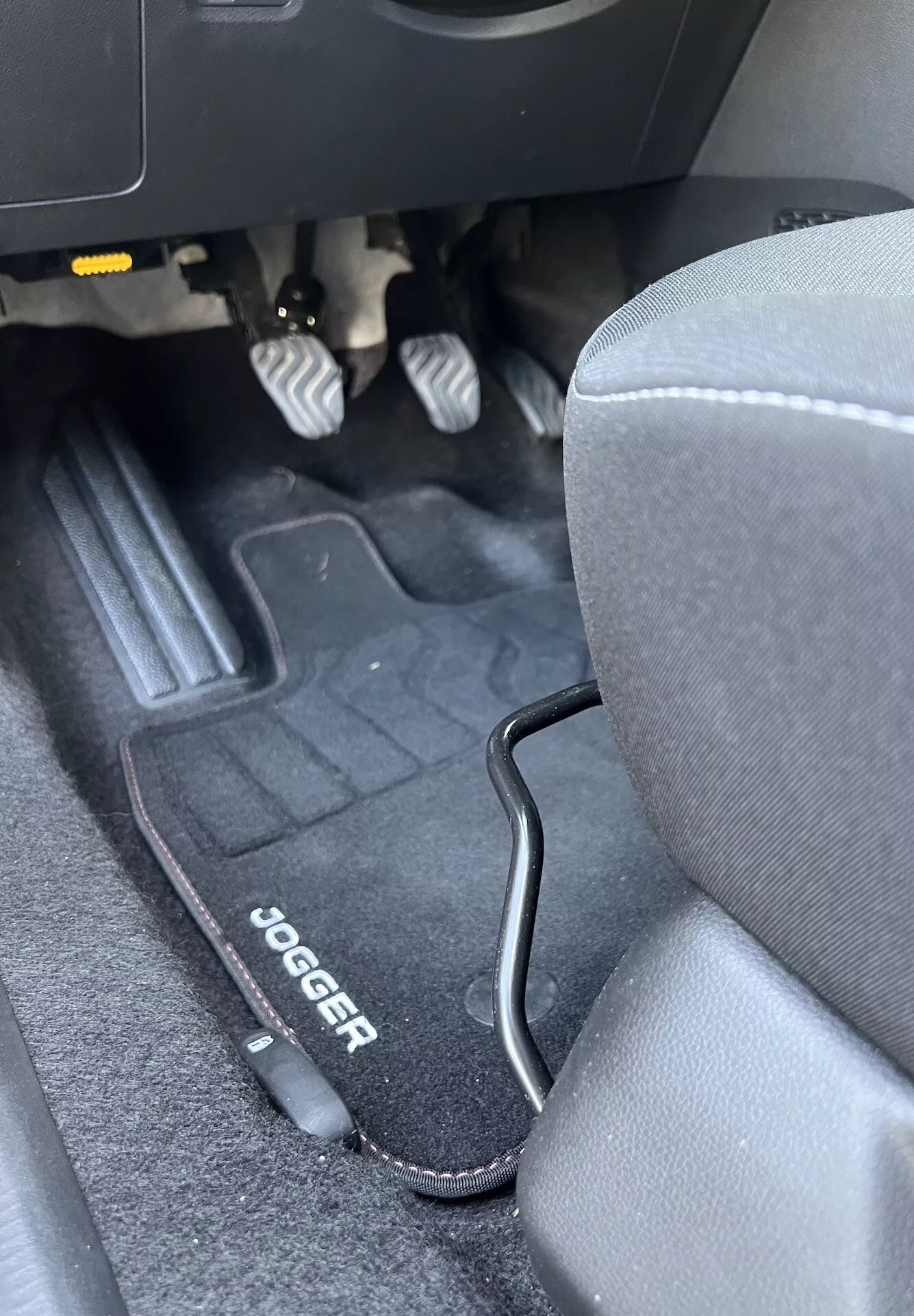Interiér Dacia Jogger 2022 ukazujúci priestor pre nohy vodiča s kovovými pedálmi, značkovou podlahovou rohožou, sedadlom zo sivej látky a čiernym plastovým lemom