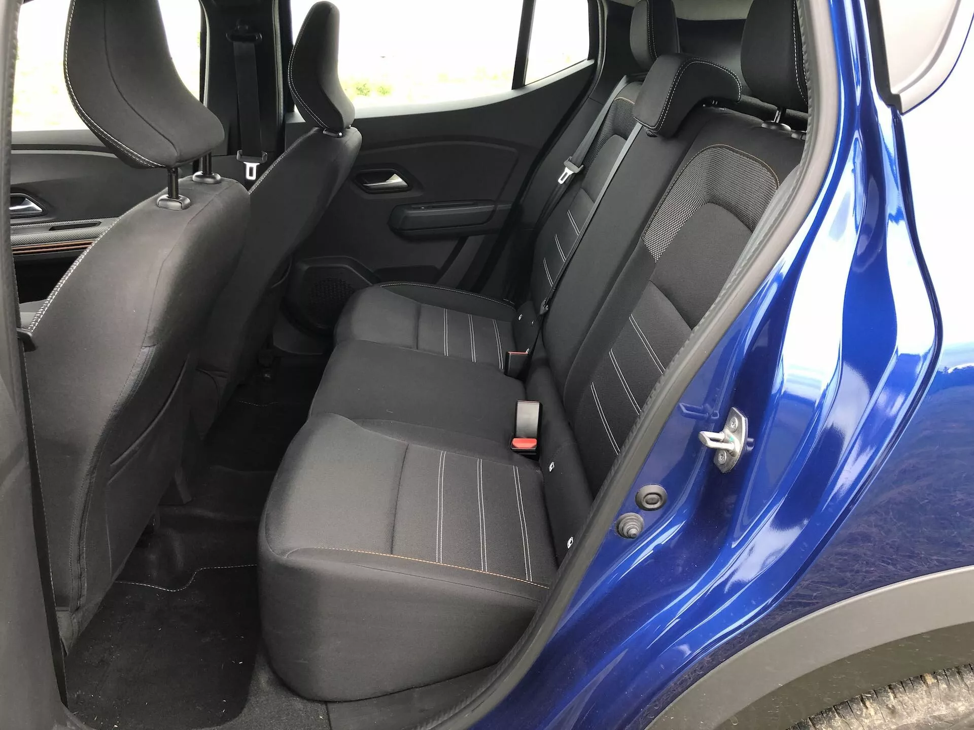 Interiér Dacia Sandero-2020 (Sandero-Stepway) so zameraním na zadné sedadlá. Pohľad mierne zhora na úroveň sedadiel, ukazujúci pravú stranu s bezpečnostnými pásmi, kľučkou na dverách a tmavým farebným prevedením s čiernym plastom a tmavošedým alebo čiernym čalúnením. Zdôrazňuje konfiguráciu sedadiel, bezpečnostné funkcie, ako sú nastaviteľné bezpečnostné pásy a možné ISOFIX úchyty, ovládanie elektrického okna na paneli dverí a celkový estetický dizajn.