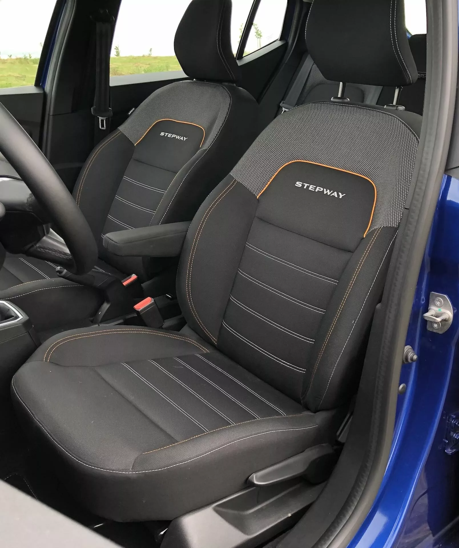 Detailný pohľad na interiér predných sedadiel Dacia Sandero 2020 Stepway, kde je vidieť vodičovo sedadlo s výraznými bočnými oporami a výšivkou 'STEPWAY' v bielej farbe s oranžovým obrysom. Sedadlá majú dvojfarebný dizajn, prevažne čierny s textúrovaným vzorom a viditeľným dvojitým šitím. Fotografia zachytáva bočné profily sedadiel z mierne zvýšeného uhla, ktorý odhaľuje hornú časť a časť sedacej oblasti, naznačujúc športovú a odolnú výbavu. Za sedadlami je viditeľná malá časť modrého vonkajšieho rámu dverí.