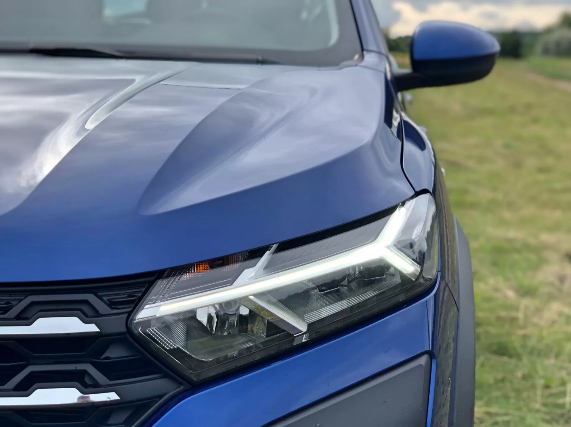 Blízky záber prednej časti tmavo modrého vozidla Dacia Sandero 2020 (Sandero Stepway) s výrazným ľavým svetlometom s dennými LED svetlami, časťou kapoty so športovými líniami a modernou prednou maskou s geometrickým vzorom. Fotografované z diagonálneho uhla mierne nad úrovňou svetiel s malou hĺbkou ostrosti, ktorá zdôrazňuje komponenty vozidla.