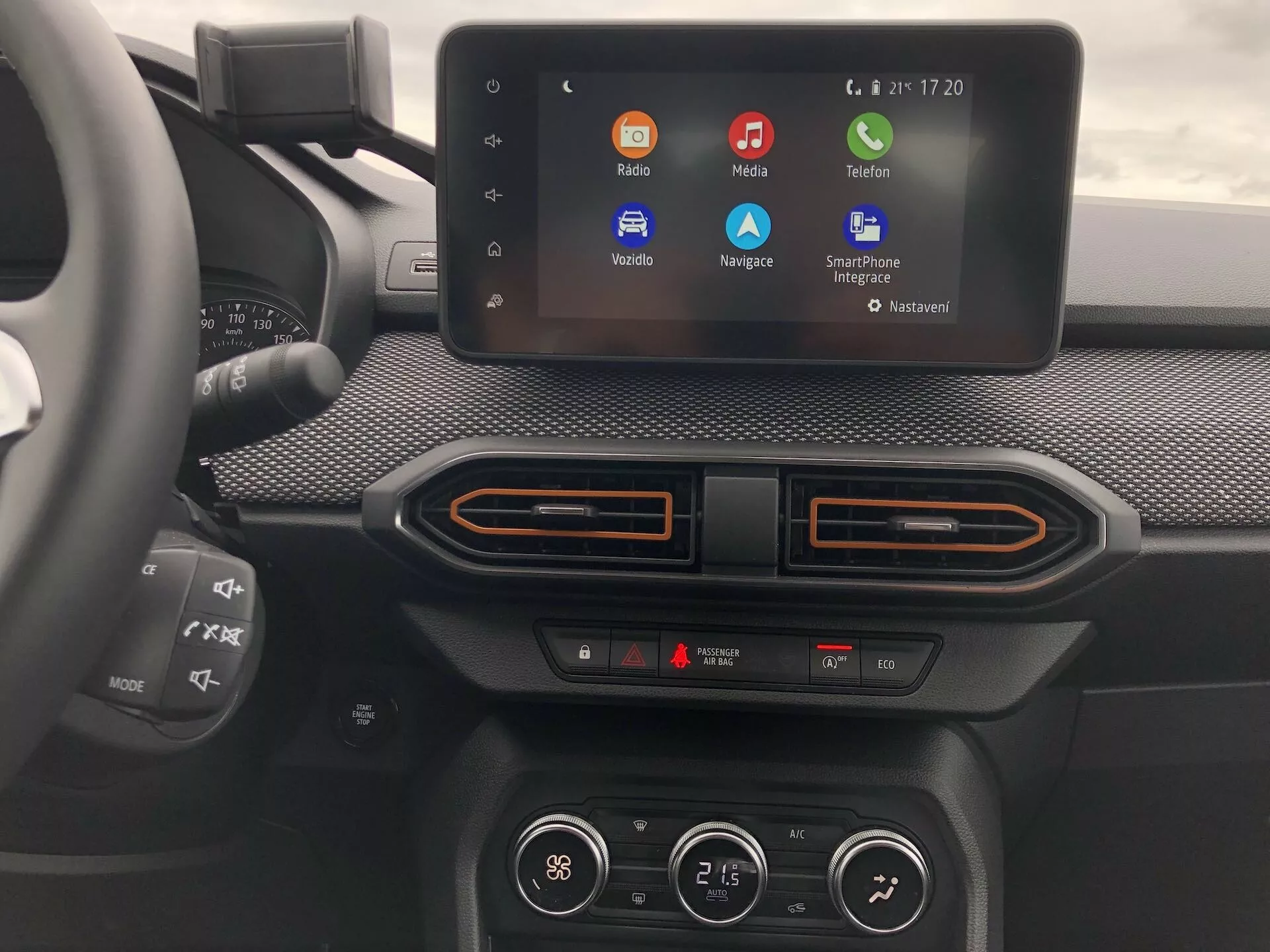 Záber na stredovú konzolu Dacia Sandero Stepway 2020, ukazujúci moderný dotykový infotainment systém s farebnými ikonami aplikácií, z perspektívy očí spolujazdca. Displej infotainmentu, umiestnený v textúrovanej prístrojovej doske so zaoblenými rohmi, naznačuje možnosti integrácie s telefónmi ako Apple CarPlay a Android Auto. Nižšie sú viditeľné ovládače dvojzónovej klimatizácie s dvoma indikátormi teploty a unikátnymi horizontálnymi ventiláciami s farebnými akcentmi. Čiastočný pohľad na multifunkčný volant vľavo. Displej zobrazuje pokročilé technologické funkcie, ako sú hlasové príkazy, navigácia a streamovanie médií.