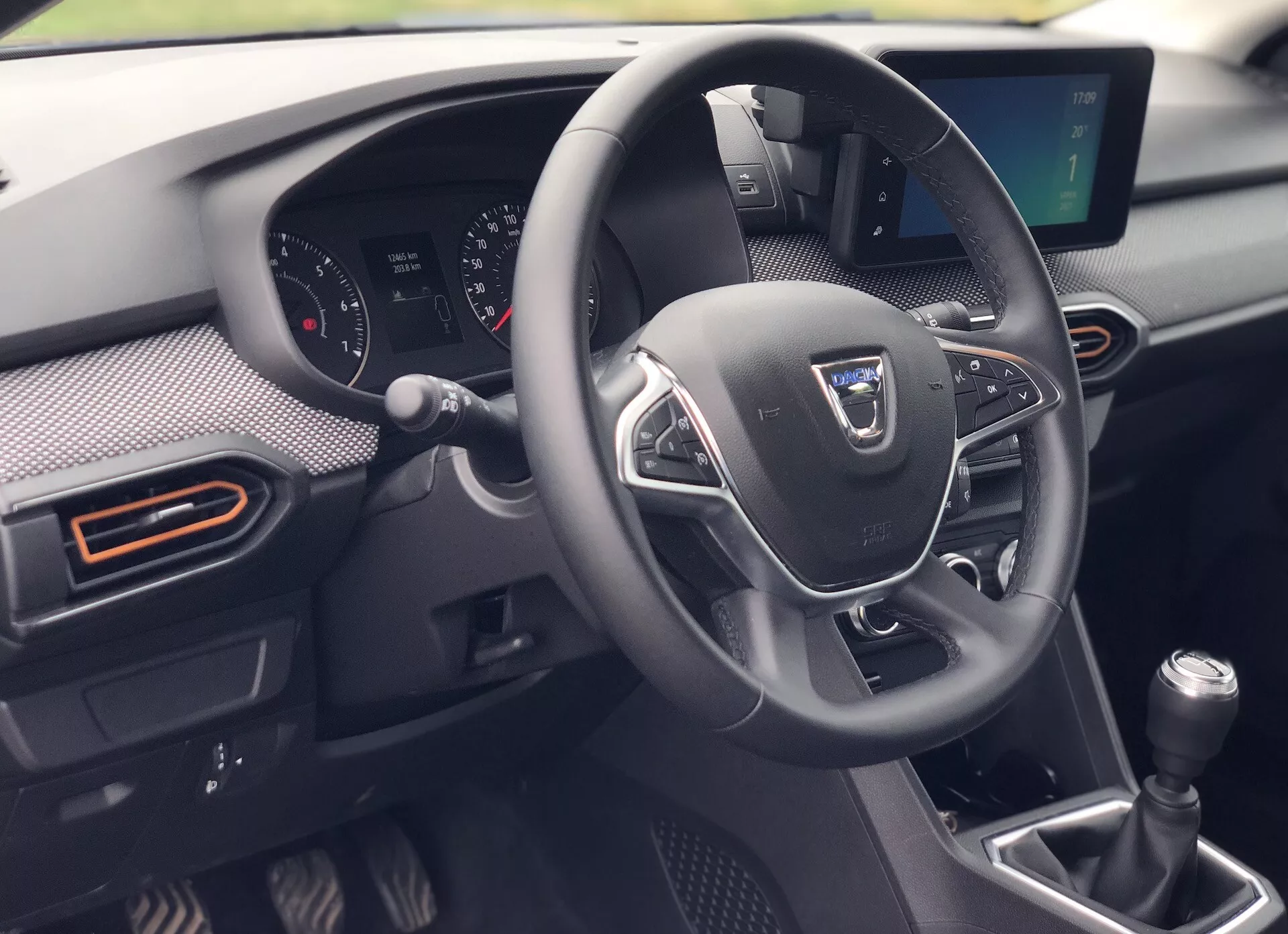 Interiér Dacia Sandero-Stepway z roku 2020 so zvýrazneným trojramenným volantom s multimediálnymi ovládačmi, čiastočným pohľadom na prístrojovú dosku s analógovými a digitálnymi displejmi, manuálnou radiacou pákou a detailným stredovým panelom s tlačidlom štart. Obrázok zdôrazňuje moderné technológie a športový dizajn s obdĺžnikovými prieduchmi s oranžovými akcentmi a obloženie v štýle uhlíkových vlákien.