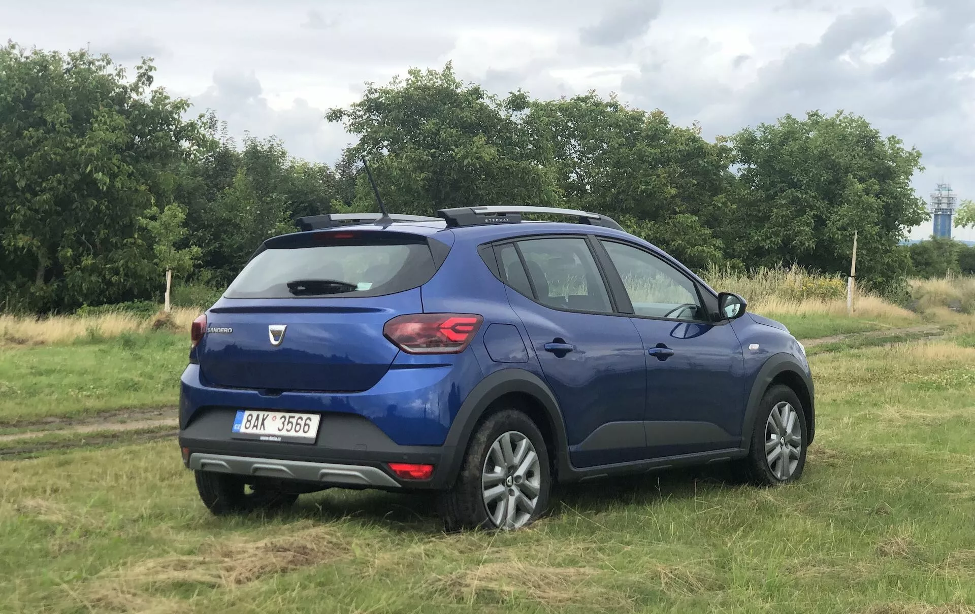 Tento obrázok zobrazuje modrý Dacia Sandero Stepway 2020, kompaktné crossover SUV, zaparkované na trávnatej ploche, zachytené z trojštvrťového zadného pohľadu. Zvýrazňuje zadné dvere so stieračom, zadné svetlá, zadný spojler, označenie značky a modelu, striebornú ochrannú dosku, dvojtónové zliatinové kolesá, strešné lišty, tónované okná a línie karosérie a kontúry vozidla.