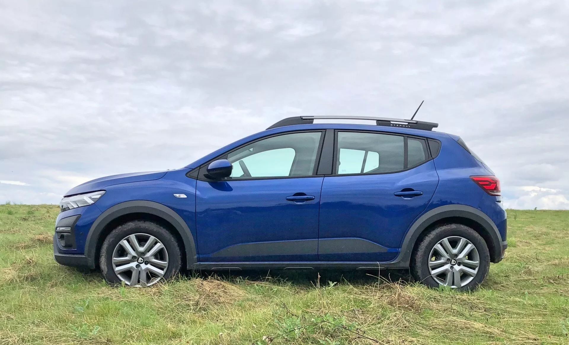 Obrázok modrého Dacia Sandero Stepway 2020, crossover SUV, zaparkovaného na otvorenom poli. Auto je zobrazené z bočného profilu, zvýrazňujúc moderný dizajn predného svetla, zliatinové kolesá a štruktúrované pneumatiky. Exteriérové prvky zahŕňajú spätné zrkadlá, kľučky dverí, charakterové línie a mierne vyvýšenie nad blatníkmi. Línia strechy sa mierne zvažuje smerom k zadnej časti a obsahuje strešné nosníky, čo pridáva na užitkovej hodnote. Vidieť aj zadné svetlo a zatmavené sklá na zadných oknách. Pozadie ukazuje mierne rozmazanú zamračenú oblohu a trávu, čo robí vozidlo hlavným objektom fotografie.