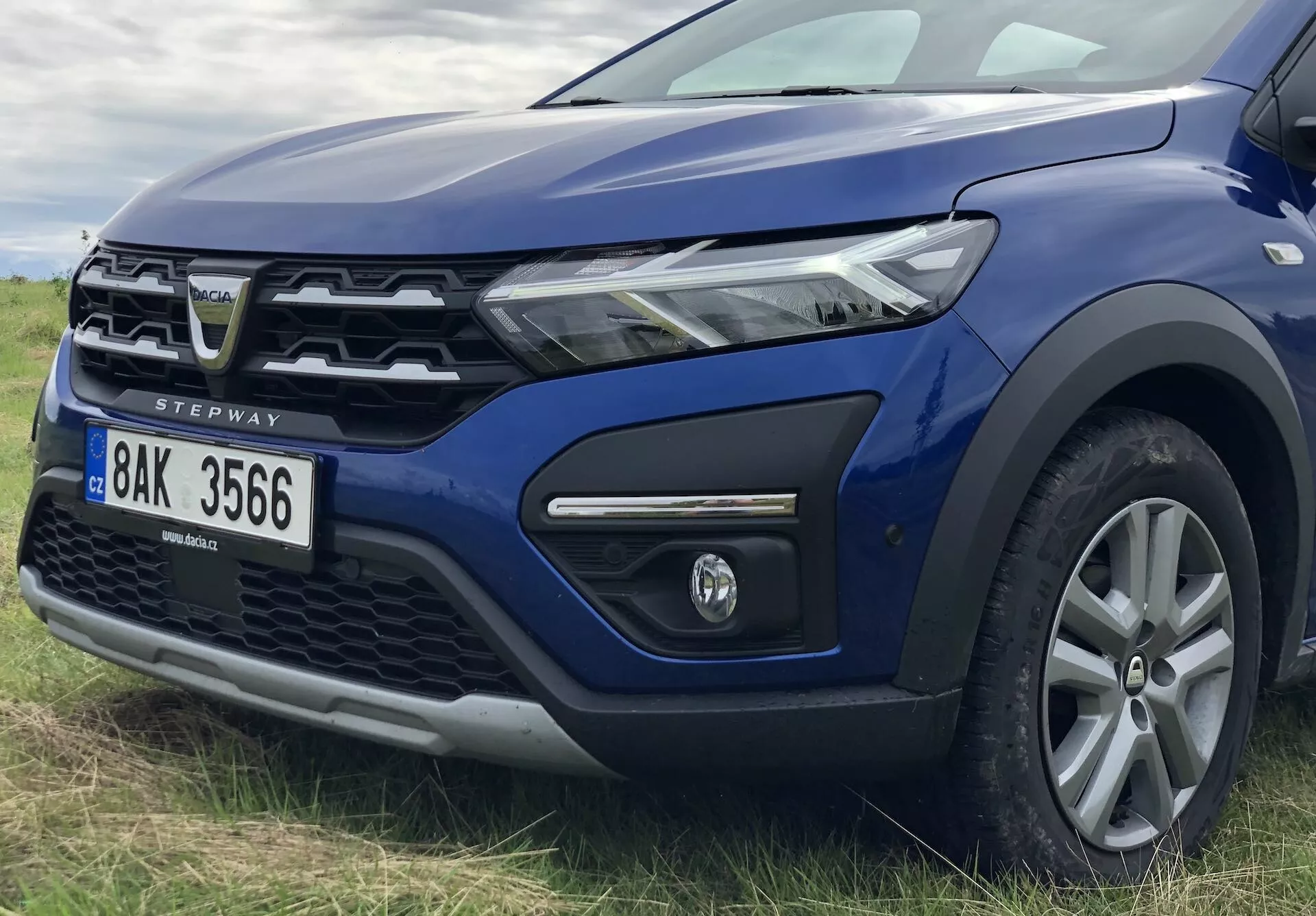Detailný pohľad na modrý Dacia Sandero Stepway 2020 z predného ľavého kvadrantu, ktorý ukazuje moderný LED navrhnutý svetlomet, čiastočne viditeľnú mriežku s charakteristickým emblémom, predný nárazník s hmlovým svetlom v čiernom ráme, zvýšenú kapotu, liatinové koleso s viacerými lúčmi, zrkadlo vo farbe karosérie, plastové obloženie na oblúk kolesa a dverí naznačujúce schopnosť terénneho jazdenia, predný sklonený čelný skla a A-stĺpik, a odznak 'STEPWAY' označujúci model.