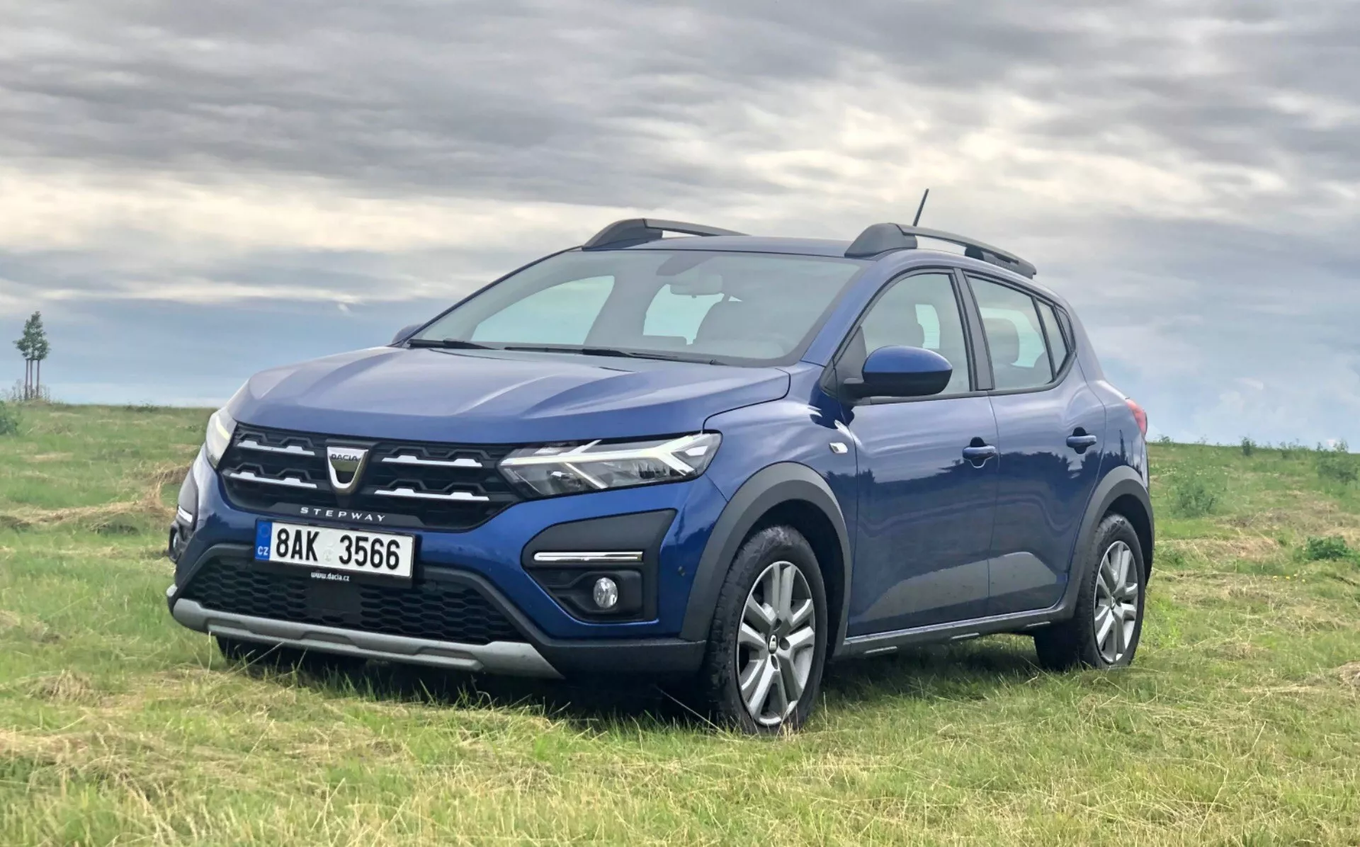 Obrázok tmavomodrého kompaktného SUV Dacia Sandero 2020 (Sandero Stepway) zaparkovaného na trávnatom poli s oblačnou oblohou v pozadí, zobrazujúceho prednú mriežku, svetlomety, predný nárazník, kolesá a profil z troch štvrtín predného uhla, mierne zvýšenej pozície.