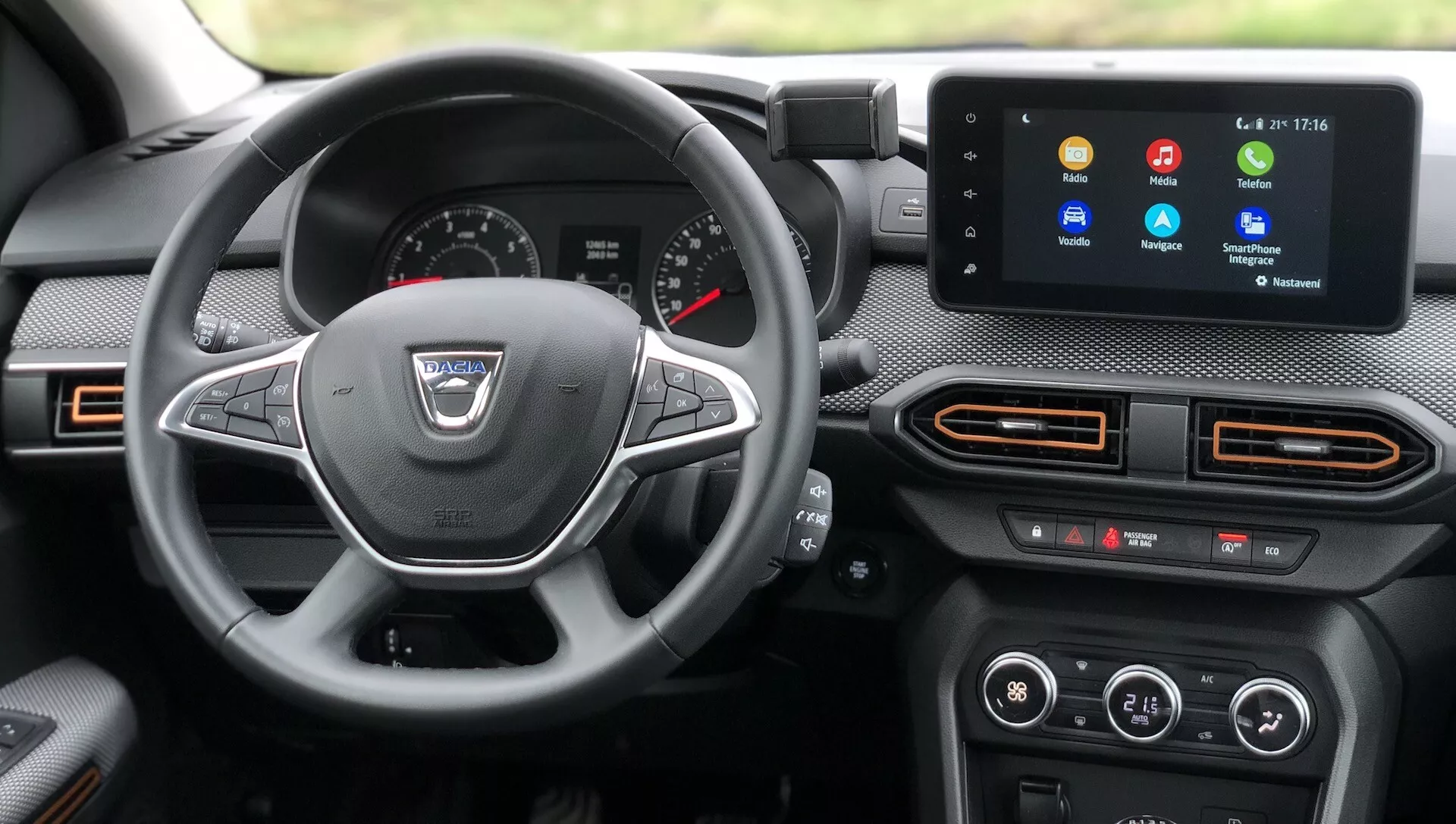 Pohľad na interiér z vodičovho sedadla Dacia Sandero 2020 (Sandero Stepway), zobrazujúci palubnú dosku, volant a centrálnu konzolu. Palubná doska obsahuje kombináciu analógových a digitálnych displejov, s dvoma veľkými analógovými ciferníkmi a menšími ukazovateľmi, vrátane centrálneho digitálneho displeja. Moderný trojramenný volant s multifunkčnými tlačidlami upozorňuje na infotainment systém. Stredová konzola zahŕňa dotykový infotainment displej s rôznymi ikonami a vzduchové výduchy s oranžovým lemovaním. Nižšie sú fyzické tlačidlá a otočné ovládače klimatizácie. Viditeľné materiály kabíny zahŕňajú kombináciu texturovaných materiálov a ambientné osvetlenie je viditeľné v oranžovej žiari okolo vzduchových výduchov.