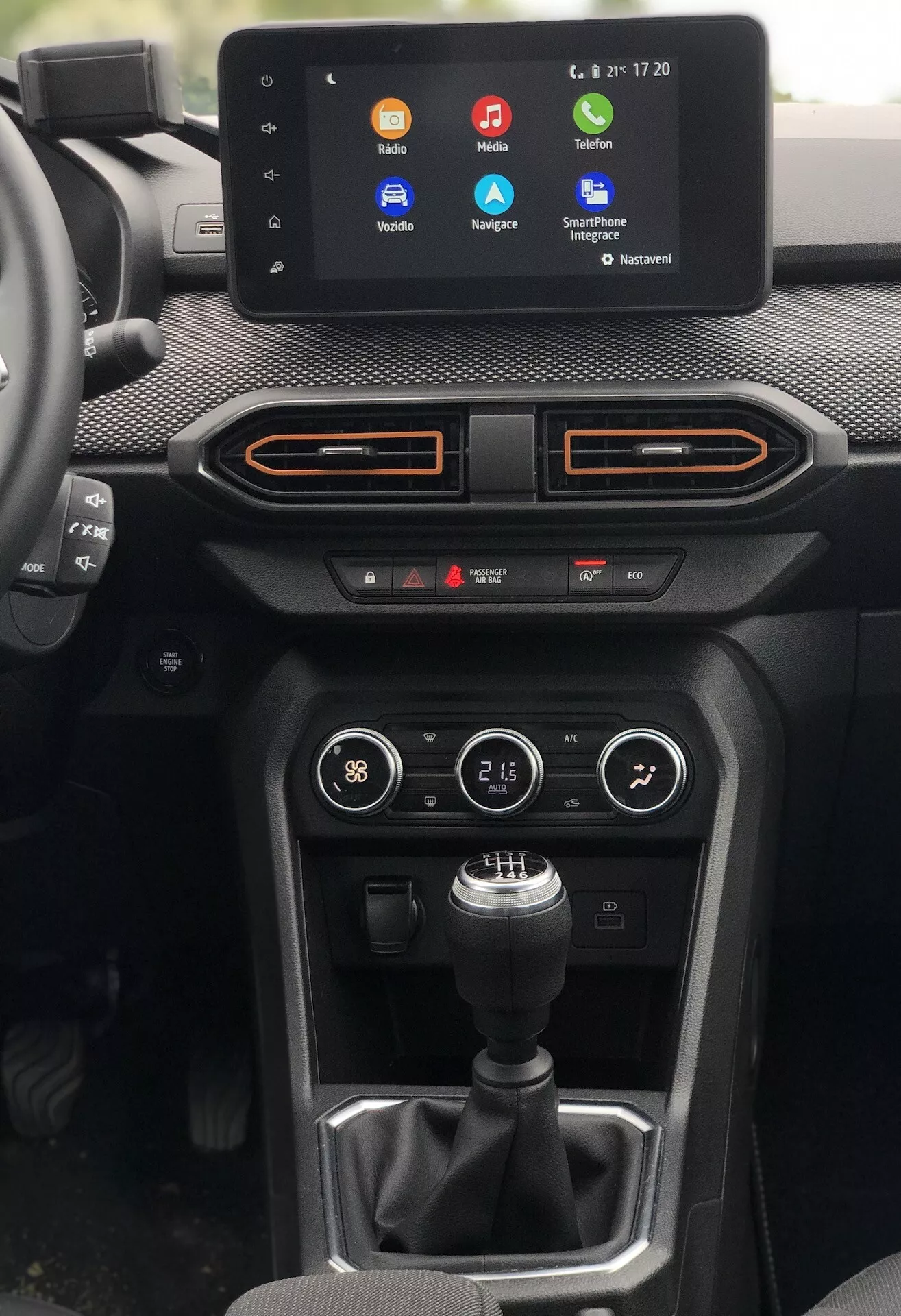 Pohľad do interiéru Dacia Sandero Stepway 2020, zameraný na stredovú konzolu z pohľadu vodiča. Významné prvky zahŕňajú infotainmentový dotykový displej zobrazujúci farebné ikony aplikácií pre hovory, navigáciu a streamovanie hudby, s časom a teplotou v pravom hornom rohu. Pod obrazovkou sú dva horizontálne prieduchy s oranžovými akcentmi, panel klimatizácie s tlačidlami a otočnými ovládačmi a manuálna radiaca páka na textúrovanom pozadí. Tlačidlo vpravo naznačuje rôzne jazdné režimy alebo ovládače a pod ovládaním klimatizácie sú viditeľné dva USB porty. Materiály zahŕňajú kombináciu mäkkého dotykového prístrojového panelu, syntetických alebo textilných textúr a plastov. Navyše, je tu ambientné osvetlenie okolo okrajov ventilačných panelov, ktoré sa zhoduje s oranžovým akcentom. Kompozícia ponúka čistý pohľad bez skreslenia, mierne naklonená doprava pre vyvážené zobrazenie.