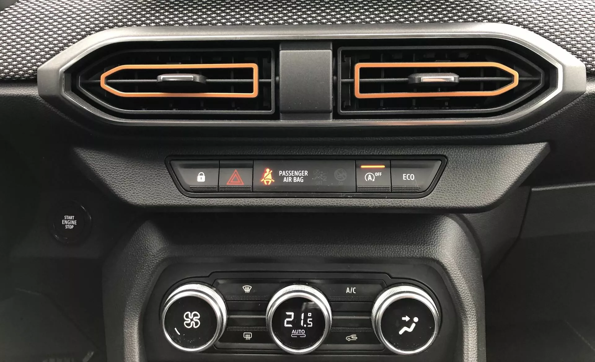 Záber zblízka na interiér auta Dacia Sandero 2020 (Sandero-Stepway), zameriavajúci sa na centrálnu oblasť prístrojovej dosky. Obrázok zobrazuje dve horizontálne orientované vzduchové vetracie otvory so vzhľadom brúseného kovu a oranžovým lemovaním, tlačidlo výstražných svetiel, indikátor stavu airbagu spolujazdca, indikátor bezpečnosti vozidla, tlačidlo eko-režimu jazdy, tlačidlo odmrazovania a klimatizačný systém s tromi rotačnými gombíkmi, ktoré zobrazujú rýchlosť ventilátora, teplotu (21,5 °C) a smer prúdenia vzduchu. Ďalšie detaily zahŕňajú textúru pripomínajúcu uhlíkové vlákno a čierne matné materiály, podsvietený displej a podsvietené tlačidlá.