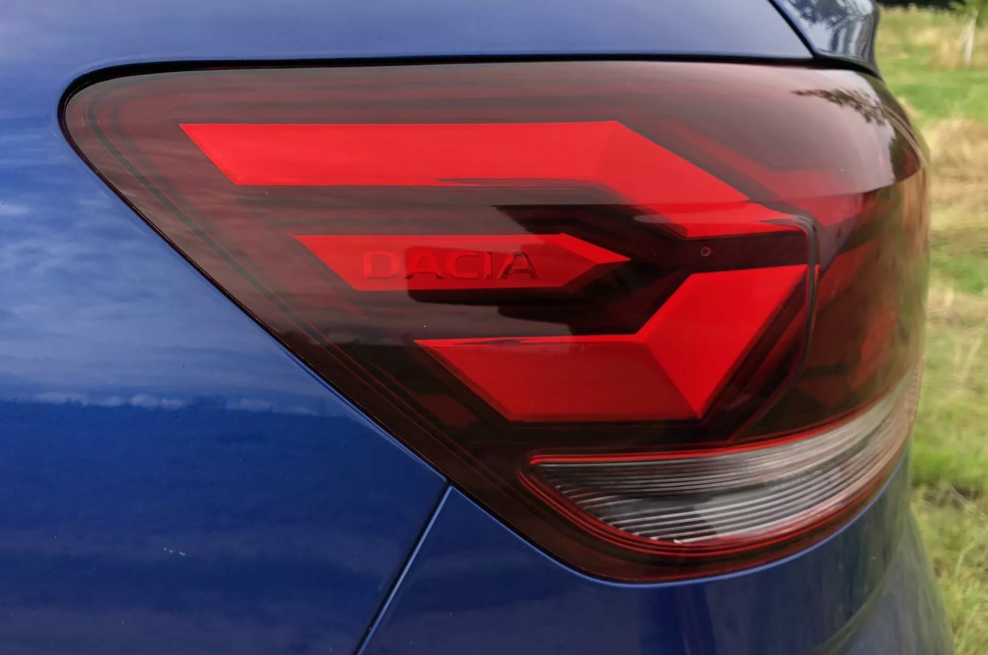 Detailný pohľad na zadnú ľavú stranu Dacia Sandero Stepway 2020 so zameraním na moderný dizajn zadného svetlometu. Zadné svetlo s uhlovými čiarami a červeným LED vzorom v tvare šípky naznačuje pohyb vpred. Jasná časť pravdepodobne obsahuje spätné svetlo a smerovku. Slovo 'DACIA' je viditeľné v obale svetla. Nad zadným svetlom je čiastočne viditeľný modrý lak karosérie.