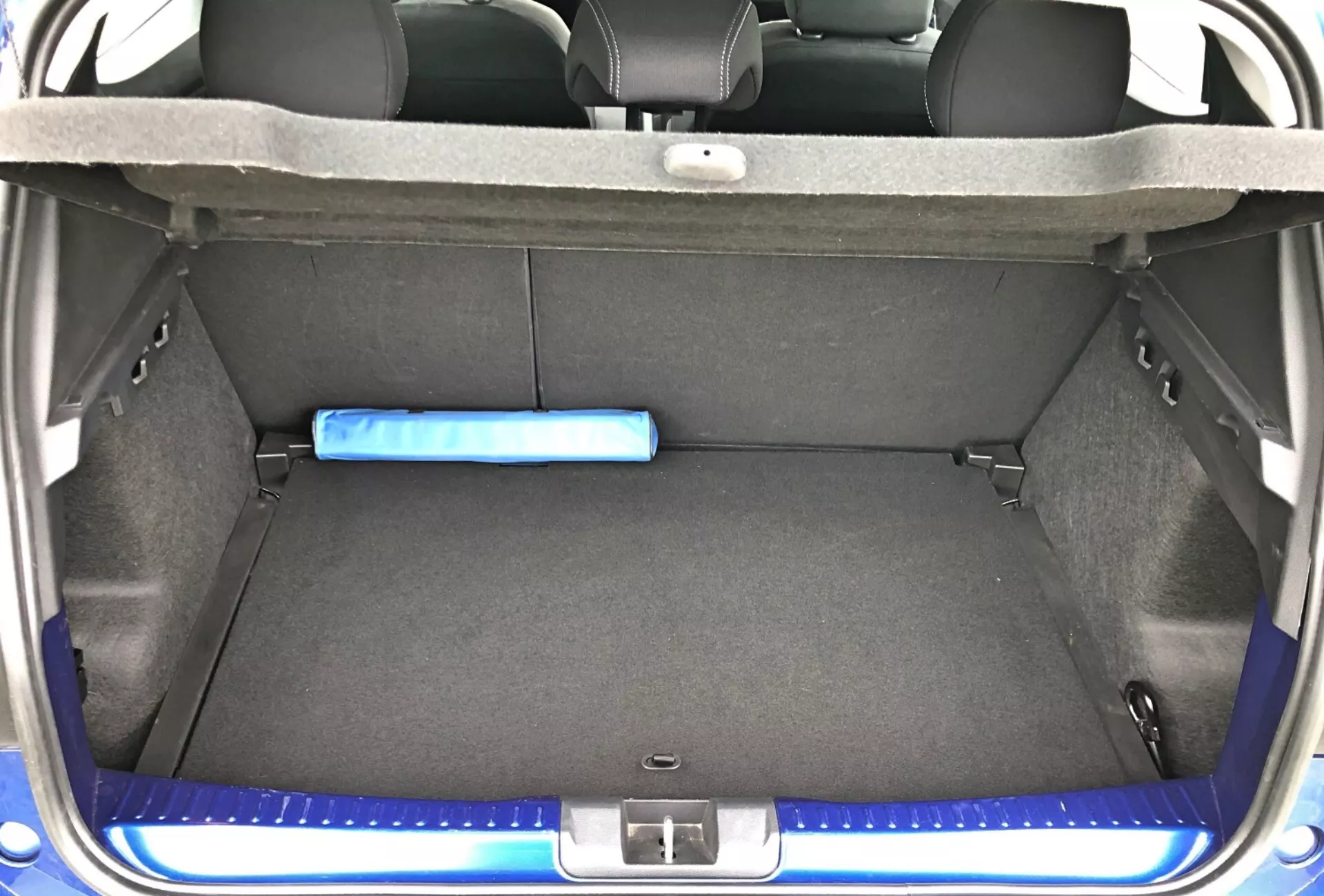 Zblízka pohľad na kufor Dacia Sandero Stepway 2020 zo zadného uhla s otvoreným kufrom, zobrazujúci čistý a prázdny priestor kufra s modrým valcovým predmetom proti operadlu zadných sedadiel delených v pomere 60/40. Kufor je vystlaný tmavošedým kobercovým materiálom a zadné sedadlá sú potiahnuté svetlošedou tkaninou. Podlaha má drážku pravdepodobne pre úložný priestor náhradného kolesa pod vyklápacou doskou. Zadný prah má ochranný modrý lem zodpovedajúci exteriéru vozidla. Viditeľná je aj malá sieťová priehradka vpravo na zaistenie malých predmetov.