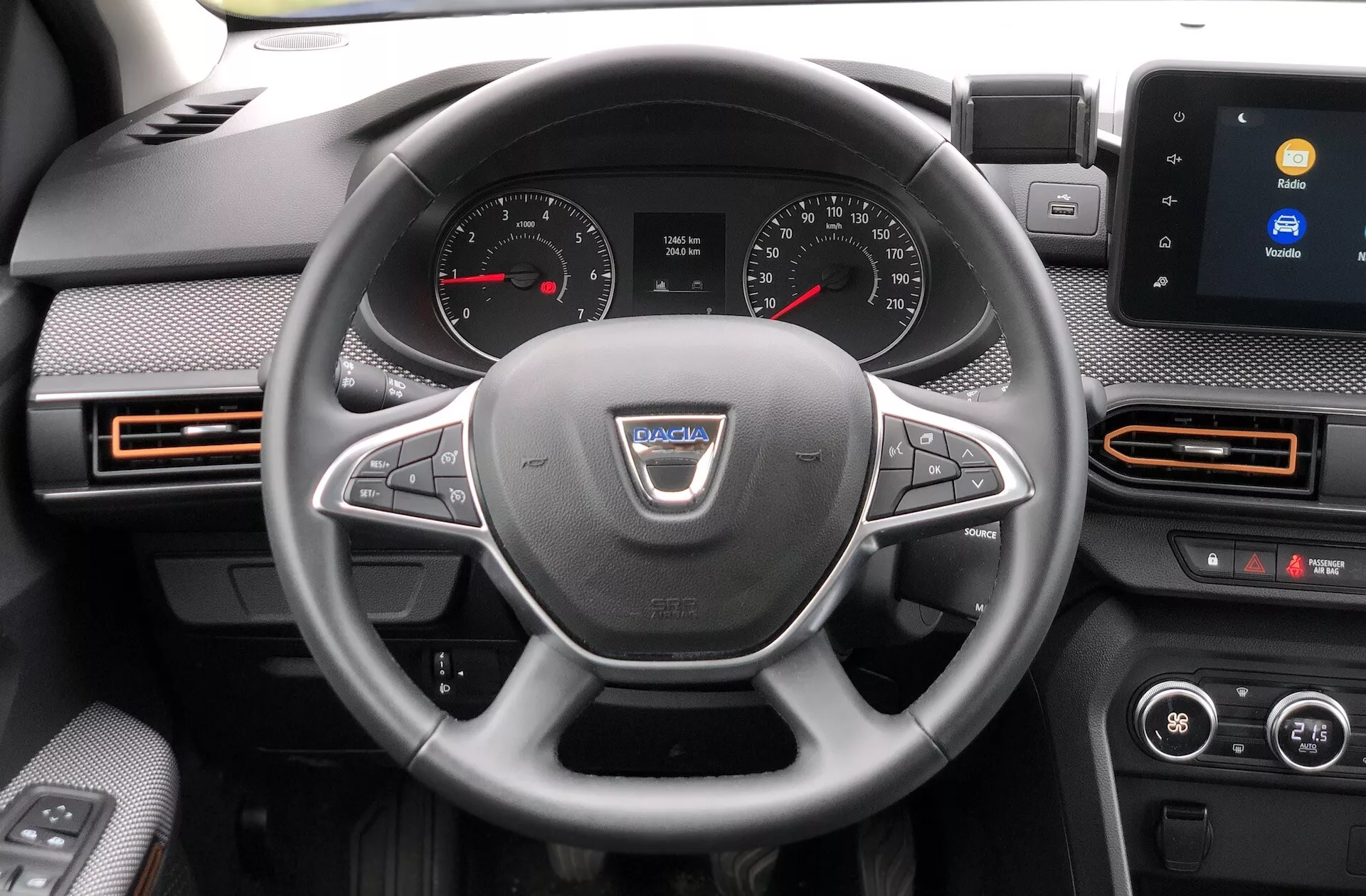 Pohľad do interiéru Dacia Sandero Stepway 2020 zameraný na palubnú dosku z pohľadu vodiča. Volant, centrálne umiestnený na fotografii, obsahuje logo výrobcu a niekoľko ovládacích tlačidiel pre funkcie ako ovládanie hlasitosti, hands-free volanie a tempomat. Za volantom je viditeľný prístrojový panel s analógovými ciferníkmi pre tachometer a otáčkomer a digitálnym displejom medzi nimi. V strede palubnej dosky sa nachádza dotykový infotainment systém zobrazujúci možnosti pripojenia, ako je Bluetooth. Viditeľné sú aj otvory s oranžovými akcentmi, dvojzónová klimatizácia a náznaky dverí vodiča s elektricky ovládanými tlačidlami okien a nastavením spätných zrkadiel.